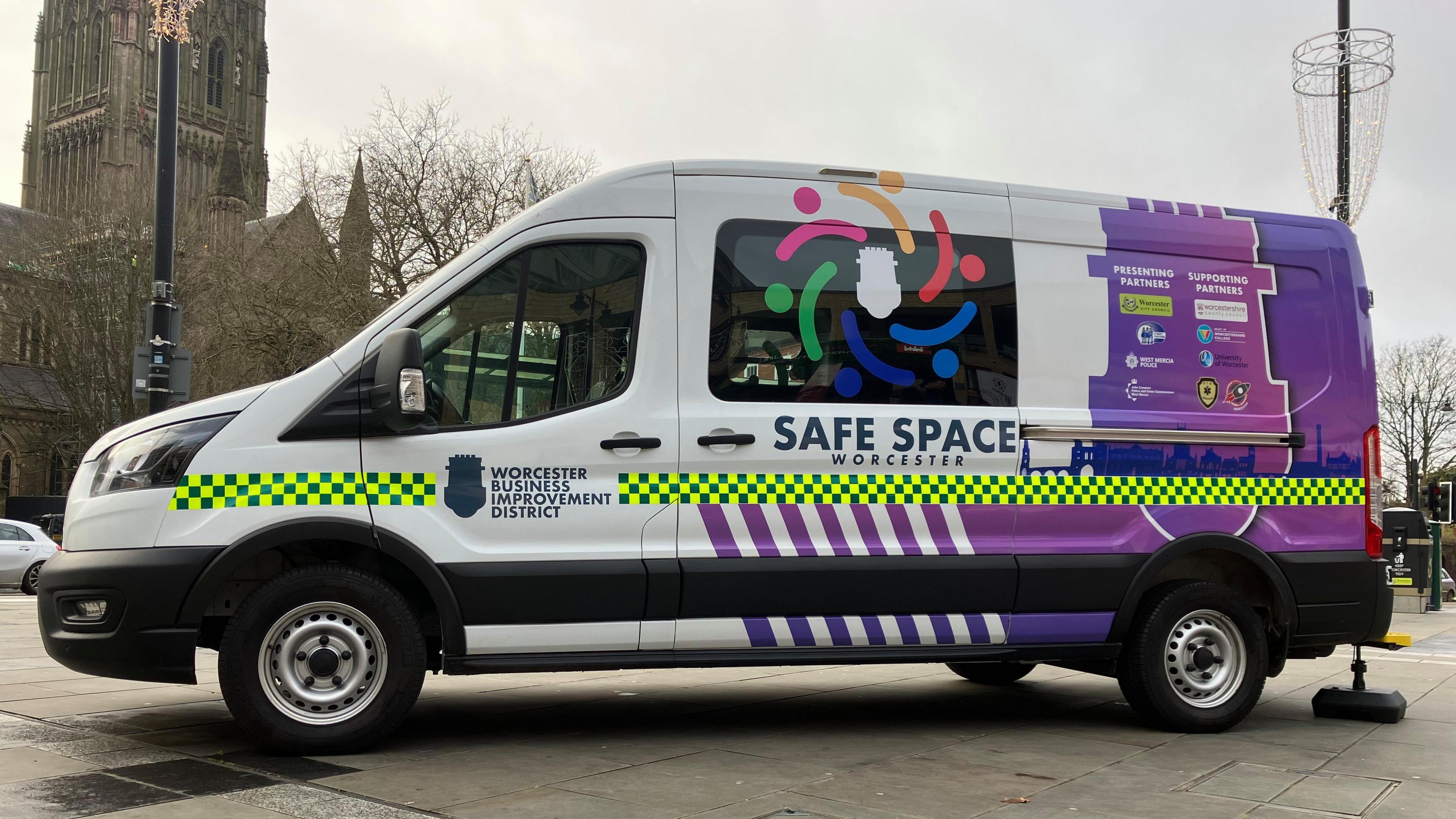 A white van with purple decals reads "Safe Space Worcester", and "Worcester Business Improvement District". It is parked on paved ground on a cloudy day, with a cathedral in the background