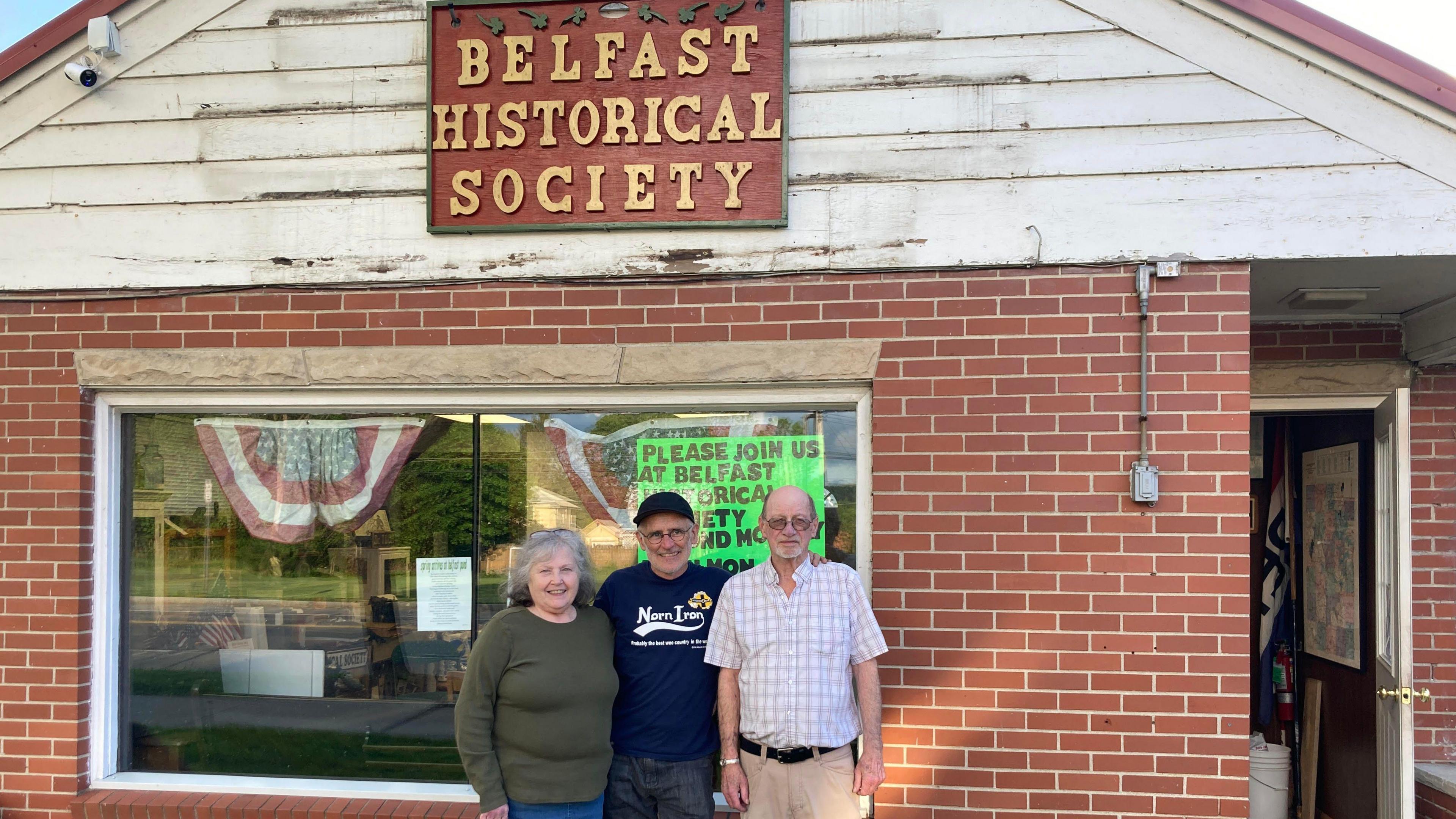 Sean Drysdale stands between a man and woman who look to be in their 50s or early 60s in front of a brick building with the sign Belfast Historical Society on it 