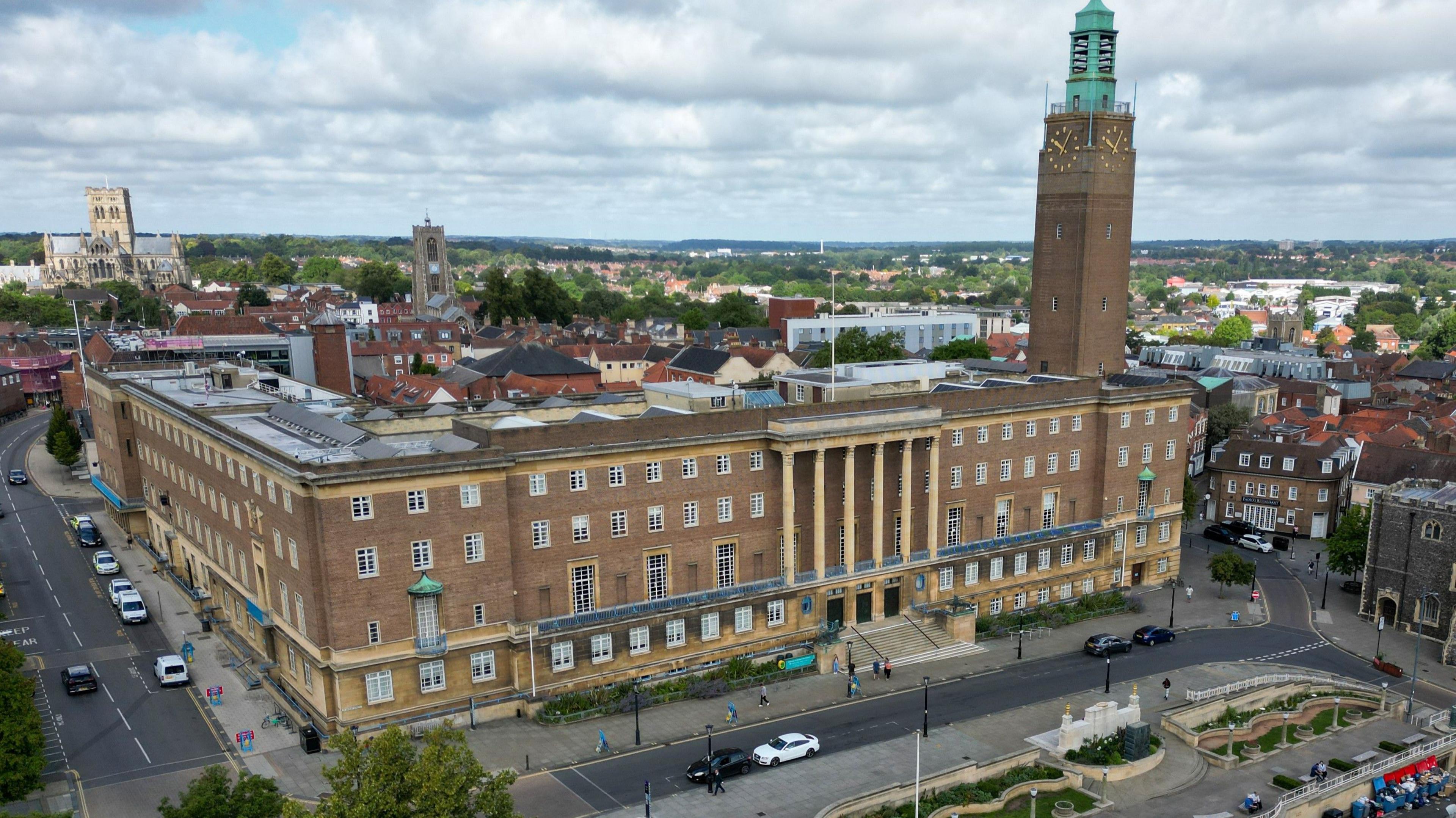 A drone image looks down on a four-storey city hall made of grey stone, featuring white windows and a balcony with columns, with a clock tower to the right of the building. It is shown surrounded by the trees and buildings of a city's urban sprawl, including a large cathedral on the horizon.