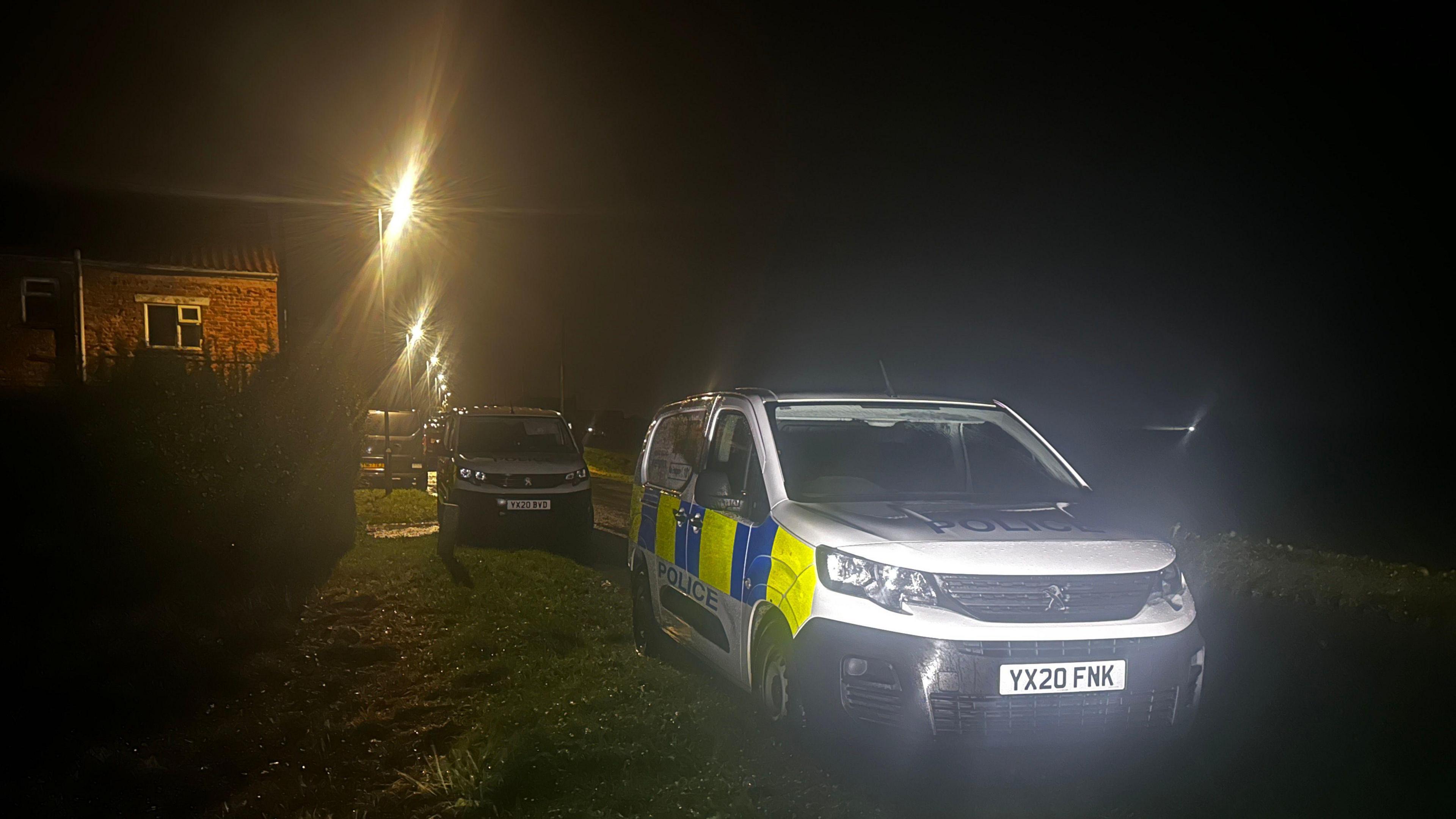 Two police vehicles are parked on a grassed verge. A red brick house is visible on the left of the image and a number of illuminated street lights are visible in the distance.