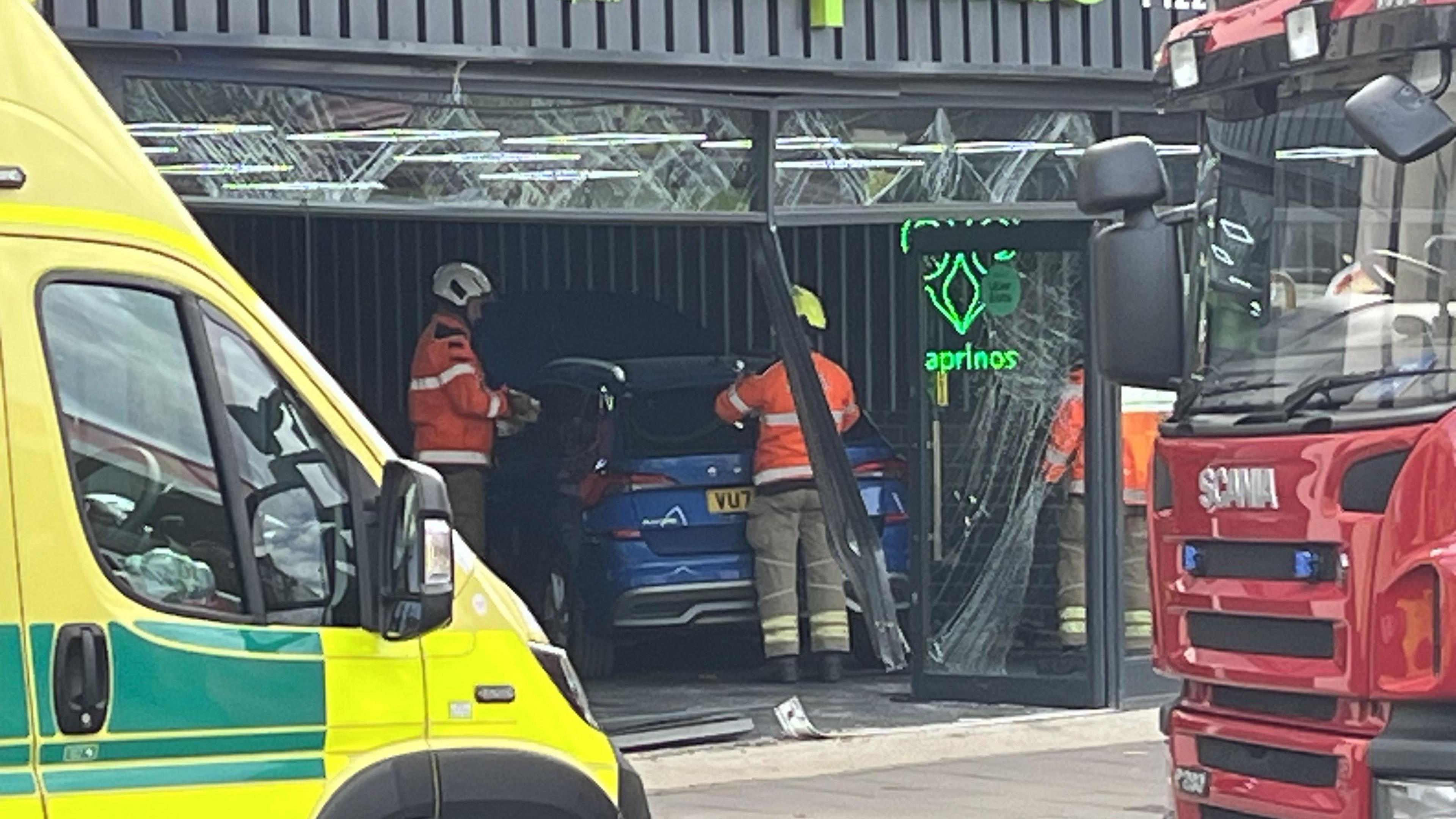 A blue Skoda inside a pizza shop with two firefighters tending to it. Outside the shop there is a fire engine on the right-hand side and a parked ambulance on the left.