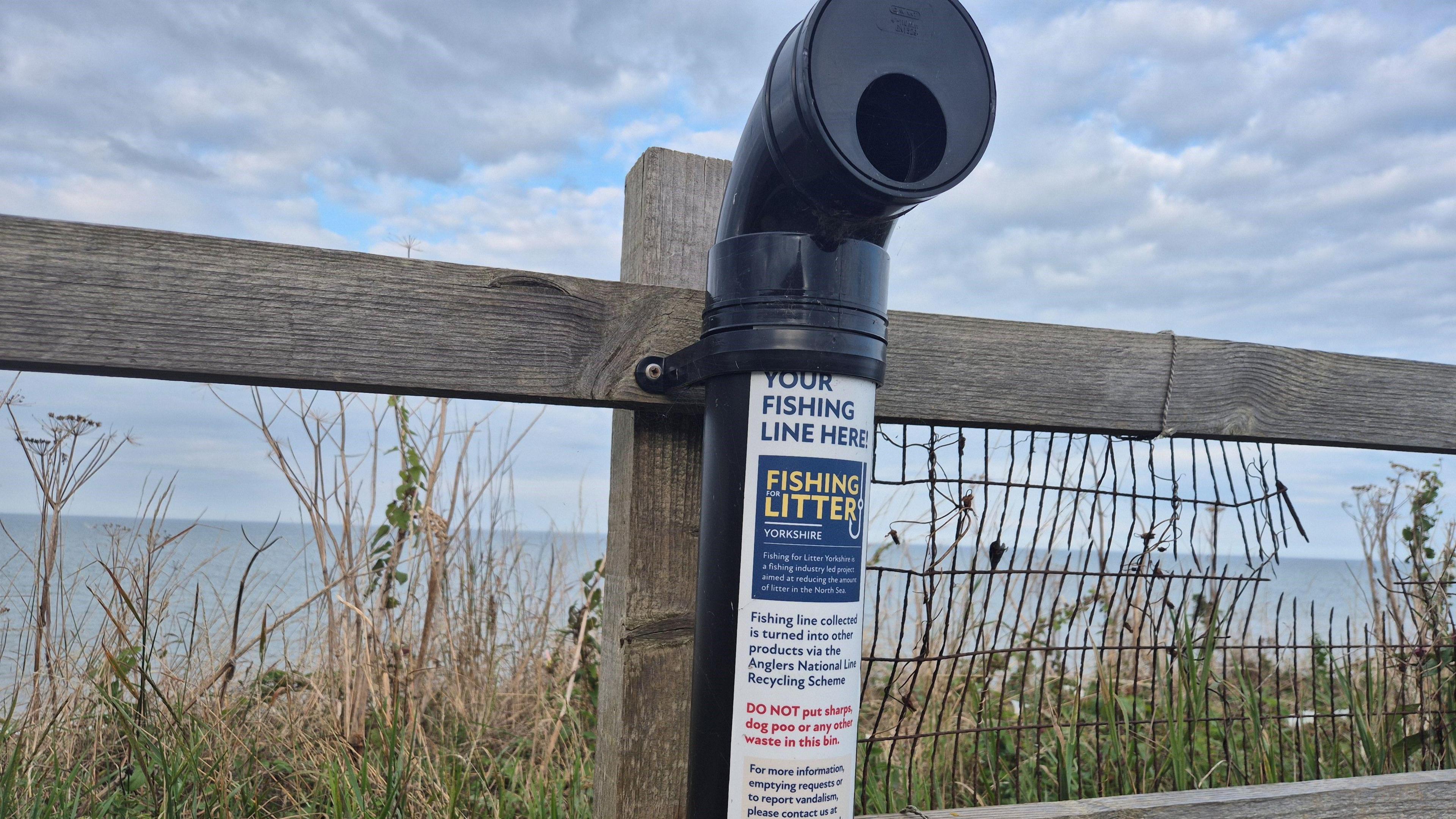 A black fish hook bin attached to a wooden fence. There is long grass around the fence.