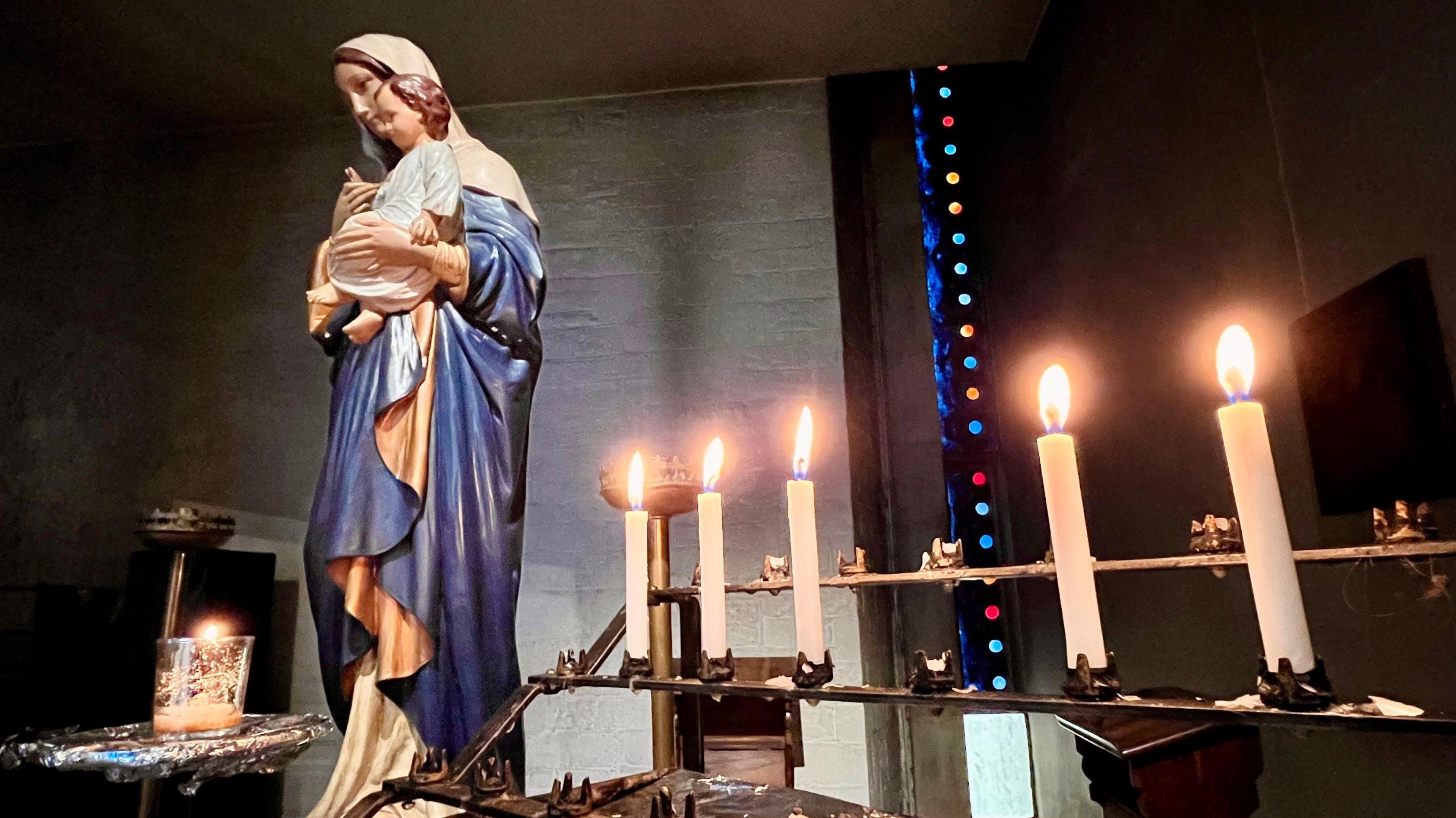 A colourful figurine of Mary holding the baby Jesus stands next to a row of candles which have all been lit inside All Saints Church in Clifton.