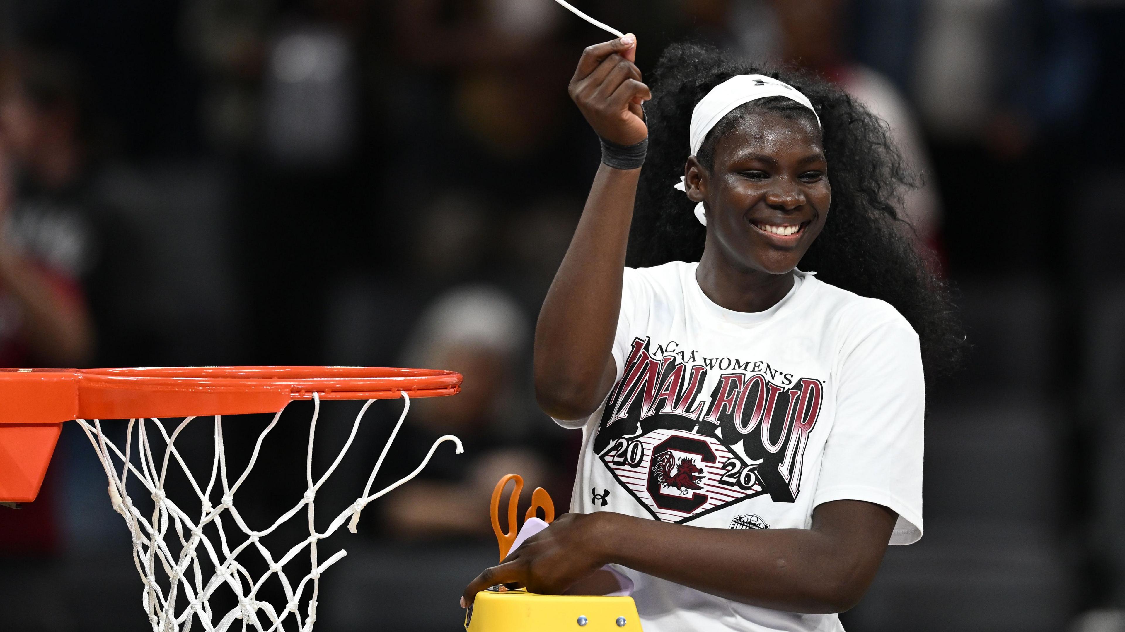 Madina Okoto, standing on a stepladder with a pair of scissors in her left hand, holds aloft a piece of basketball net which she has just cut off a basketball hoop next to her. She is wearing a predominantly white t-shirt and white headband, and is beaming with a smile
