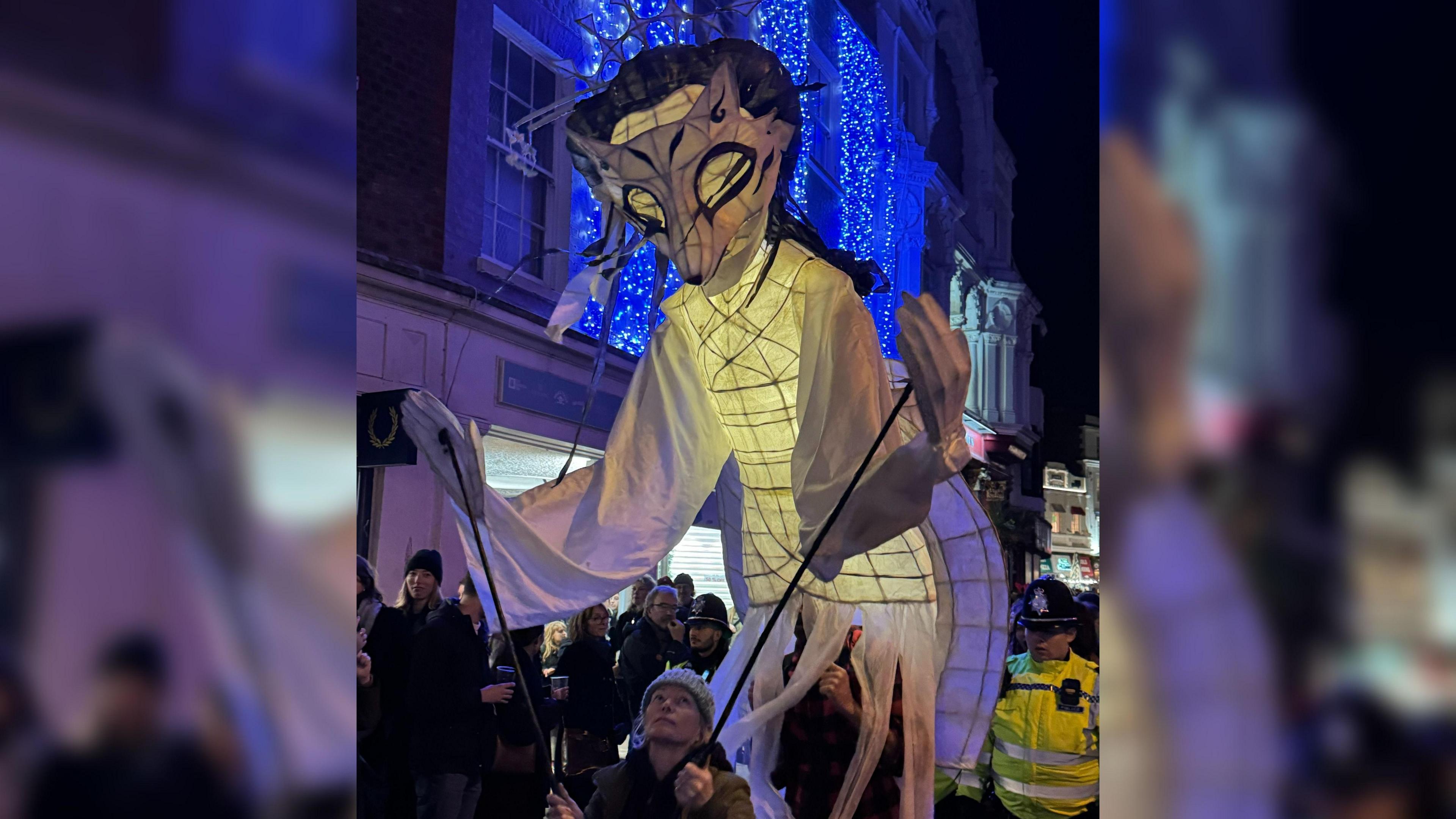 A lantern in the shape of what looks like a fox with its front paws controlled by two sticks seen held by a person standing just in front of it. There are fairy lights strung on a building in the background and people lining the streets