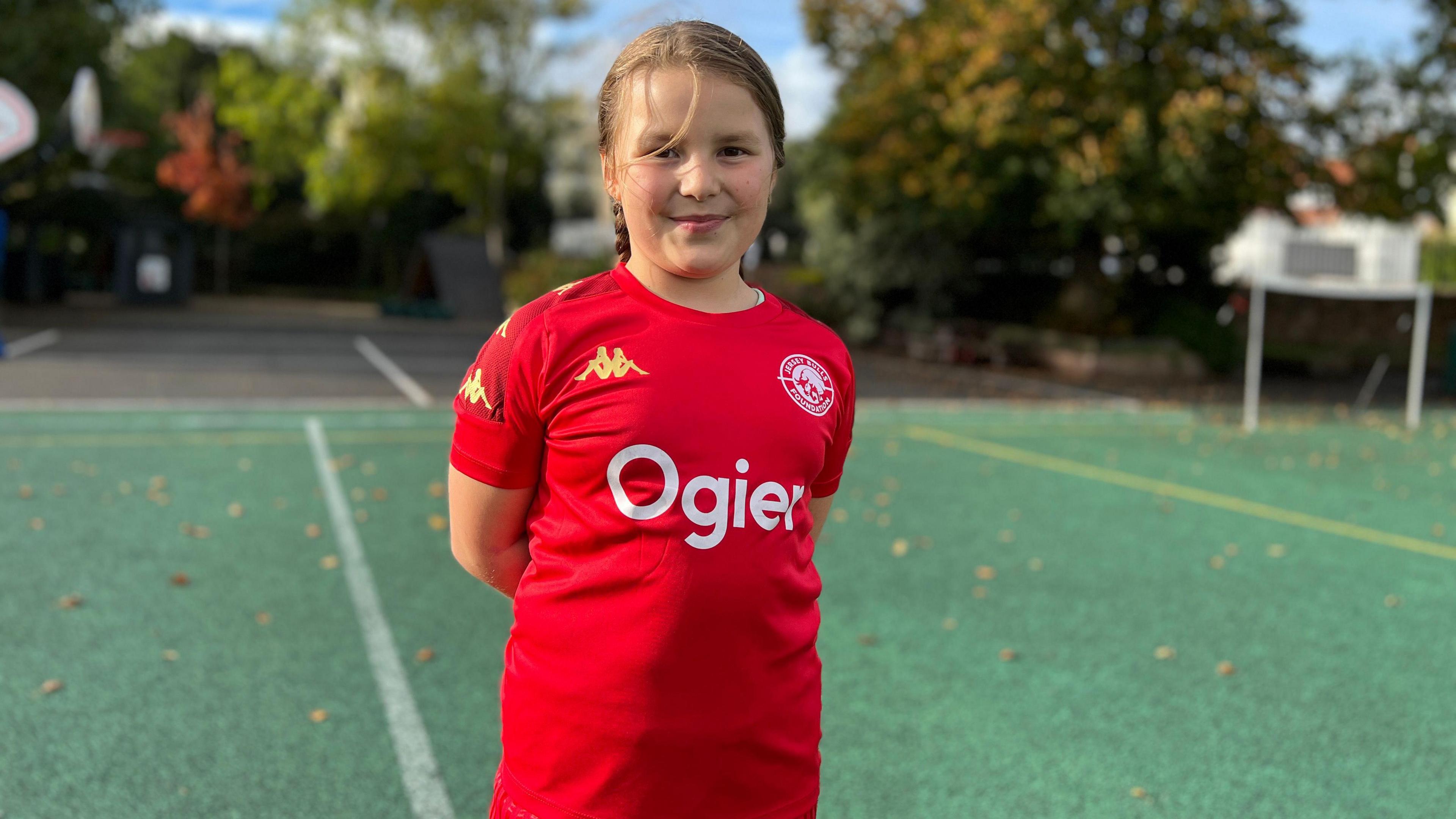 Emelia smiles at the camera with her hands behind her back. She's wearing the red Jersey Bulls Foundation training top which has Ogier on it and a white logo of the foundation on her chest. Behind her is the artificial pitch at the school's playground with trees in the distance.