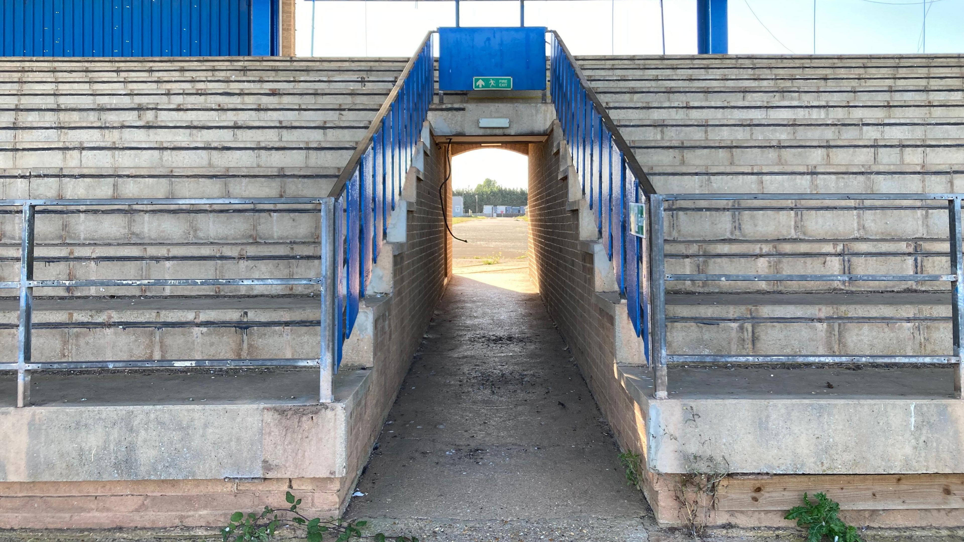 An image of the speedway grandstand. The seats have been taken out so there are concrete steps and a gangway. There are weeds in the bottom of the photo.