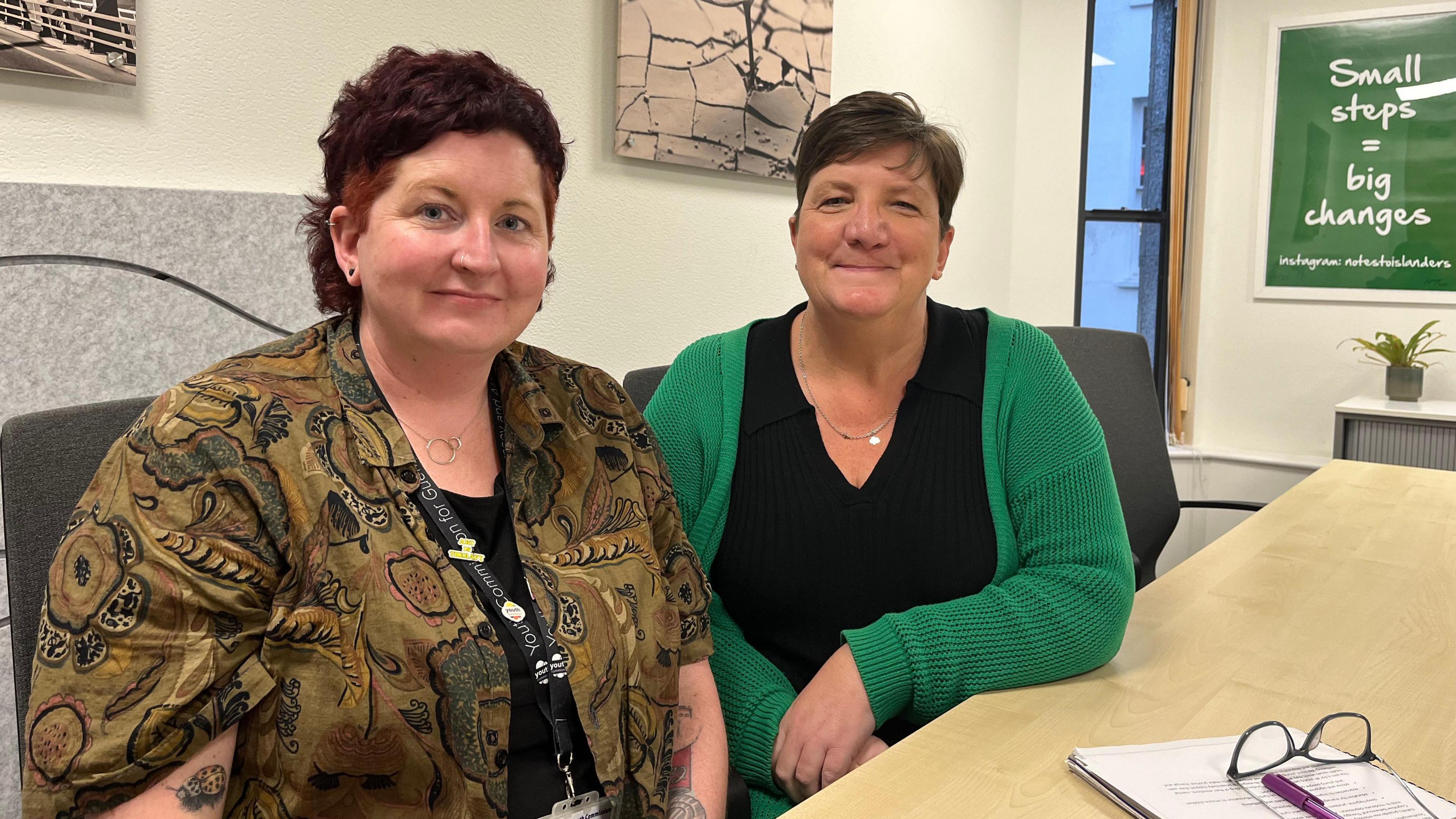 Two women sitting at a table, smiling at a camera. One is wearing a green patterned shirt and one is wearing a black T-shirt and a green cardigan.