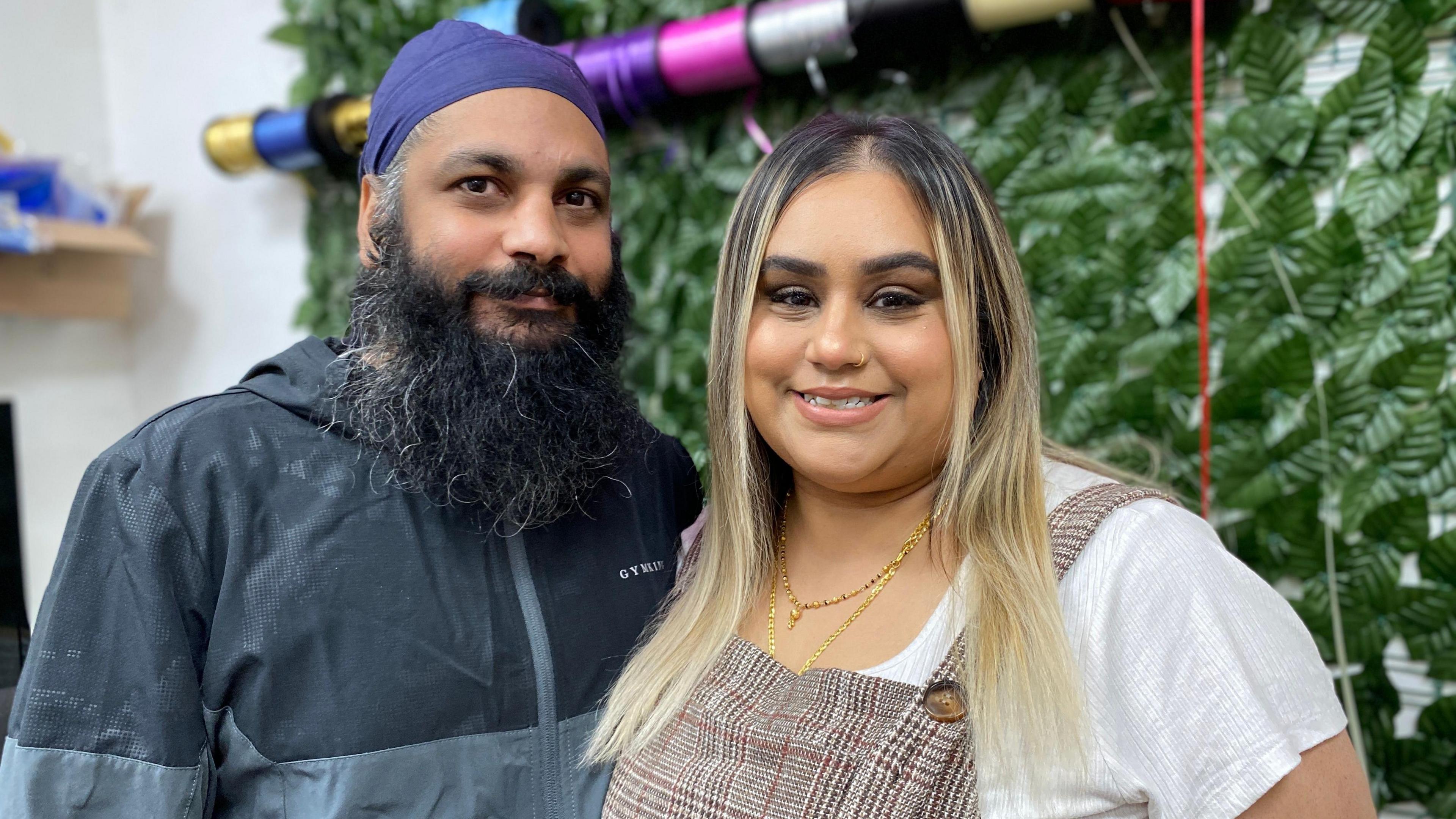 A man and a woman are smiling together. The woman has blonde hair and is wearing a white top and a plaid pinafore. The man has a dark-coloured beard and is wearing a blue turban and a black jacket.