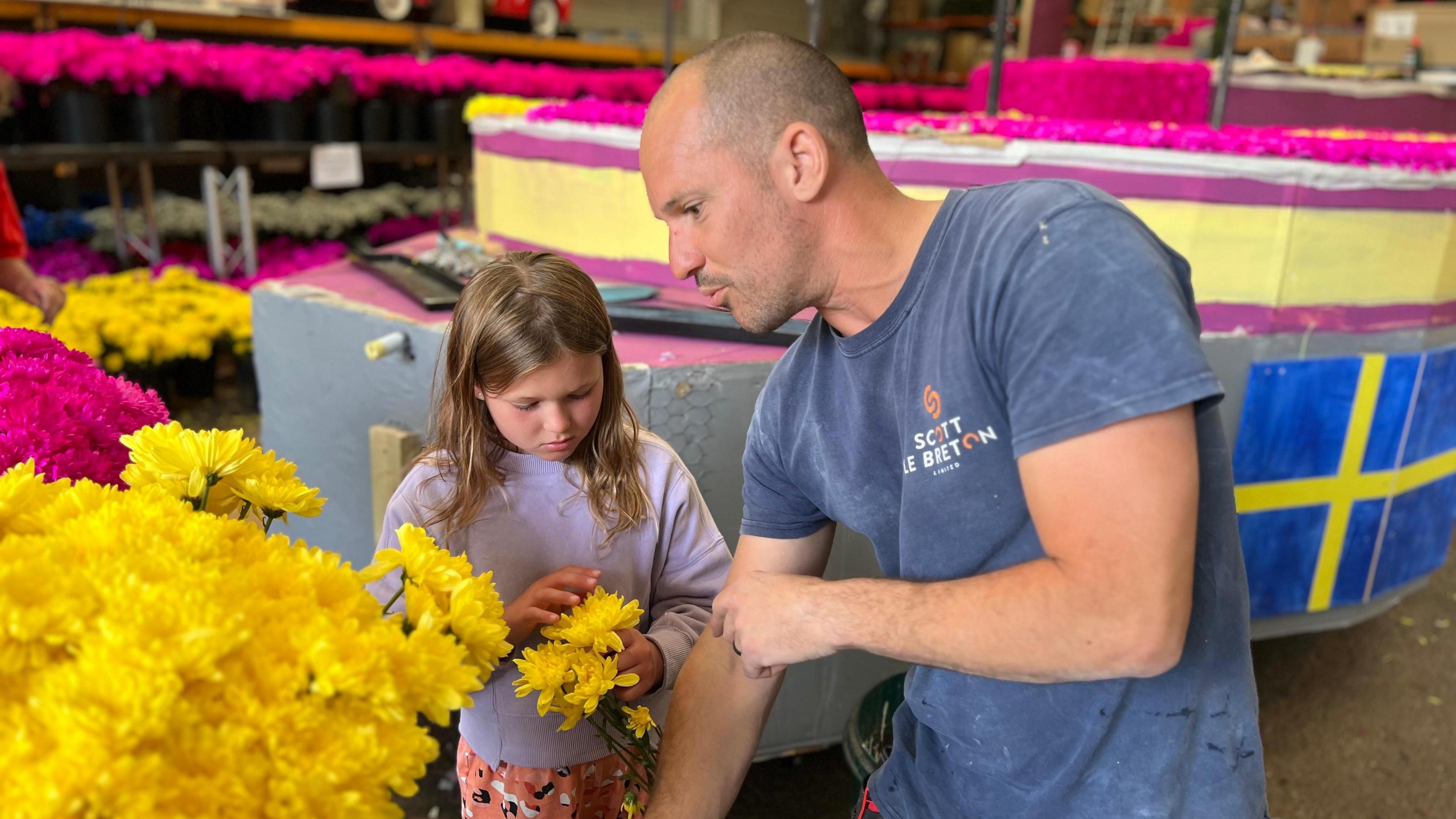 A father and daughter looking at a bunch of yellow flowers in front of the St Peter unfinished Eurovision float, there are pink and yellow flowers surrounding the float