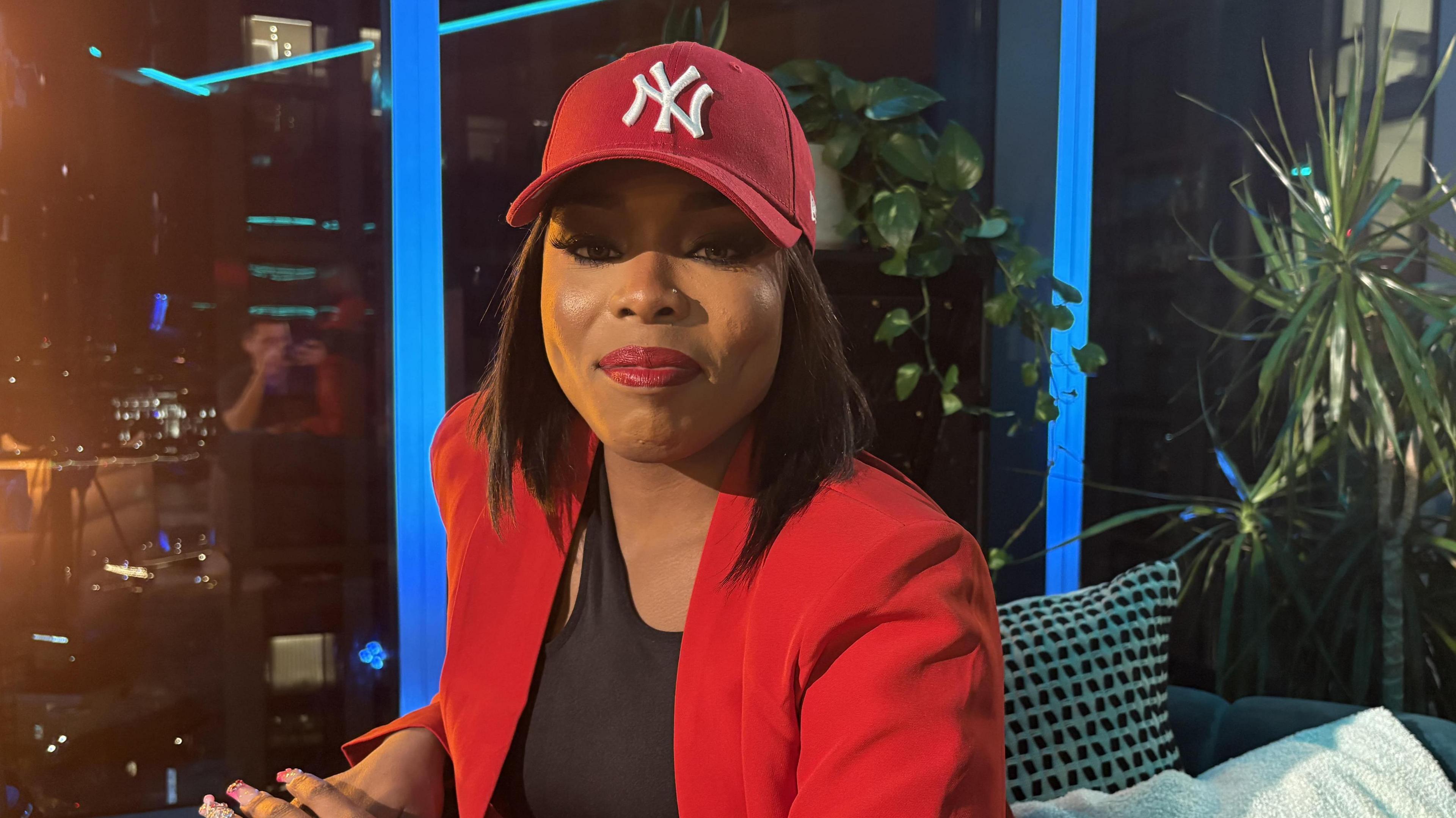 Layyah on a sofa with her hands on the side. Wearing a red blazer and a red New York Yankees cap. 