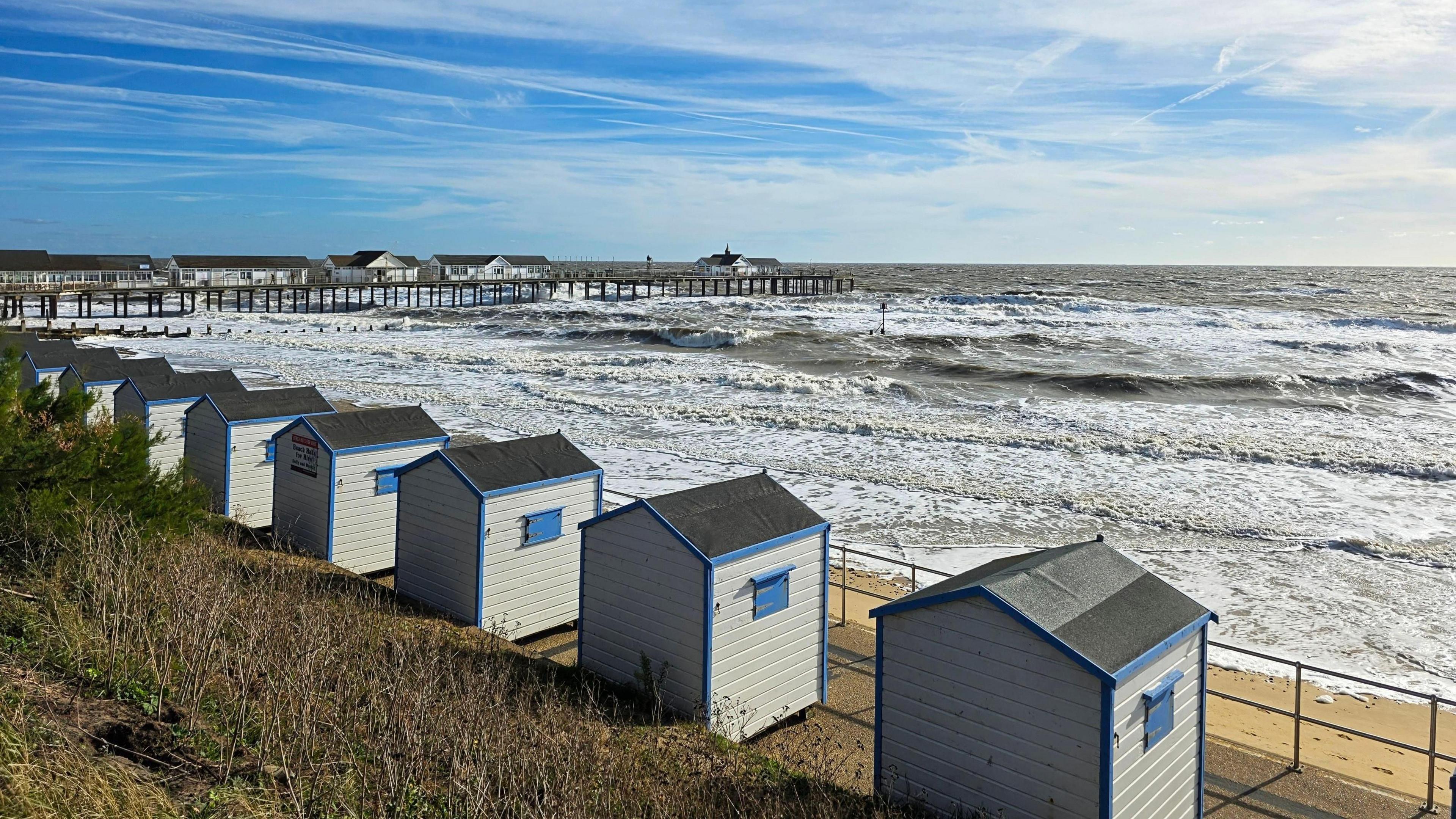 The coast at Southwold during a windy day, with waves turning white as they break. Beach huts rest on a hill above the sea. A pier is to the left extending out into the sea. 