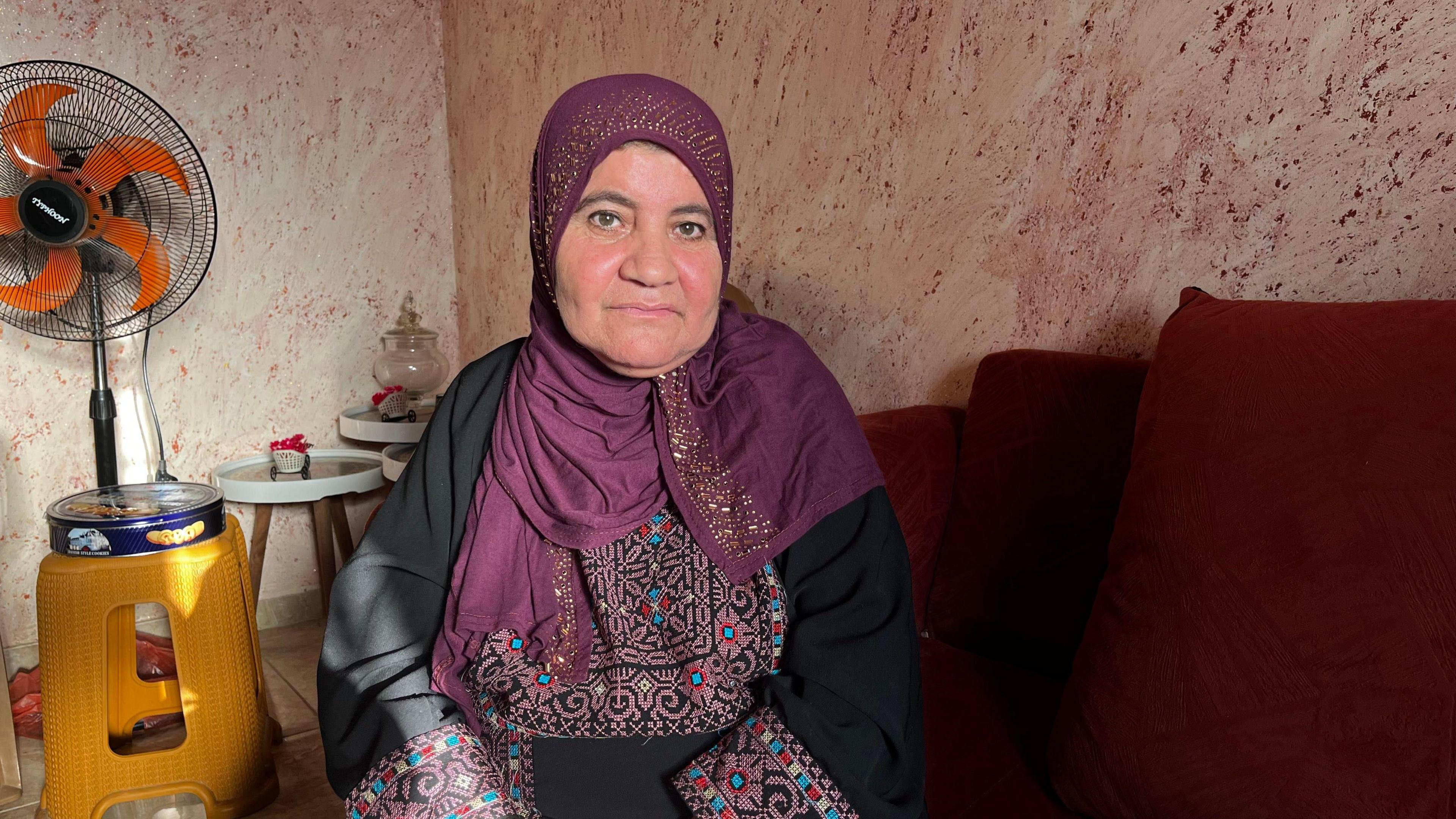 Afaf abu Alia. She is wearing a black dress with patterns around the sleeves and on the chest. She is wearing a deep red heascarf with gold detailing. She is sitting on a red sofa, the wall behind her is beige with a red pattern and there is a fan next to some tables nearby
