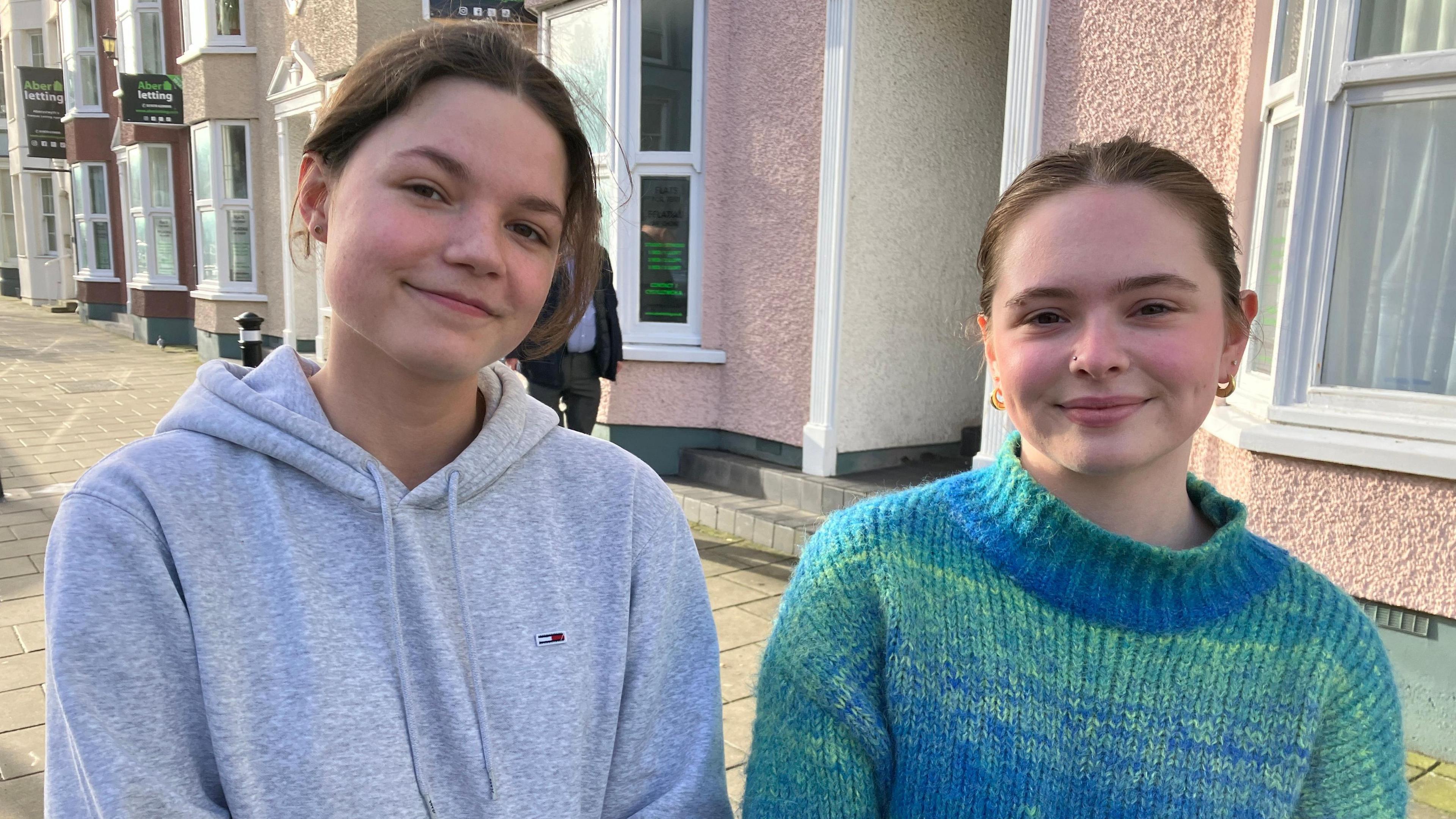 Two girls pictured in a sunny residential street in Aberystwyth. Poppy (left) wears a grey hoodie and has her brown hair pulled back behind her ears. Molly (right) wears a green and blue knit turtleneck jumper and gold hoop earrings with her brown hair tied back. 