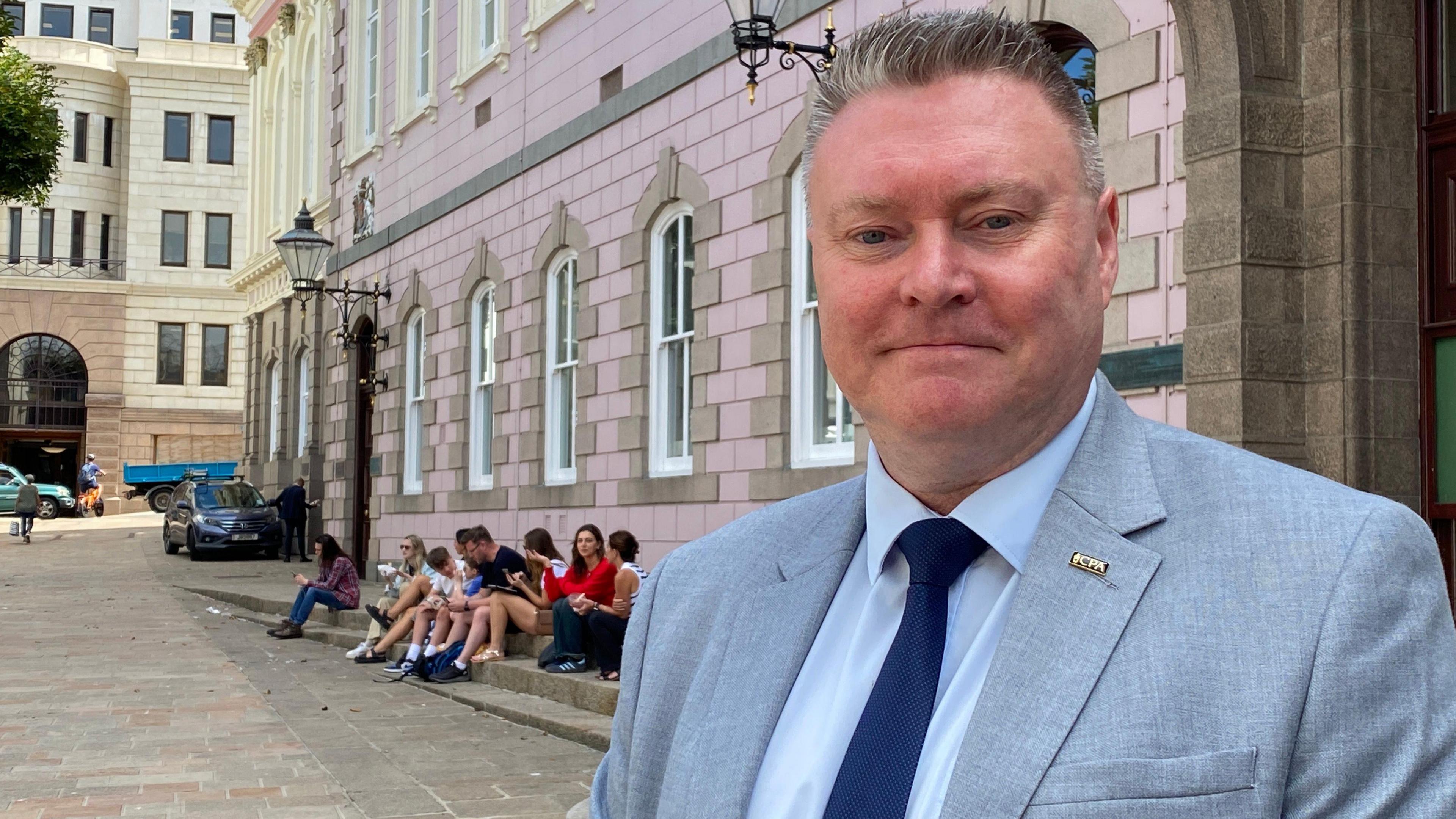 A picture of Deputy Rob Ward who is a man with short spiky grey hair. He is wearing a light blue suit with a pale blue shirt and dark blue tie. He is stood to the right hand side which a row of children sat on steps behind his shoulder.