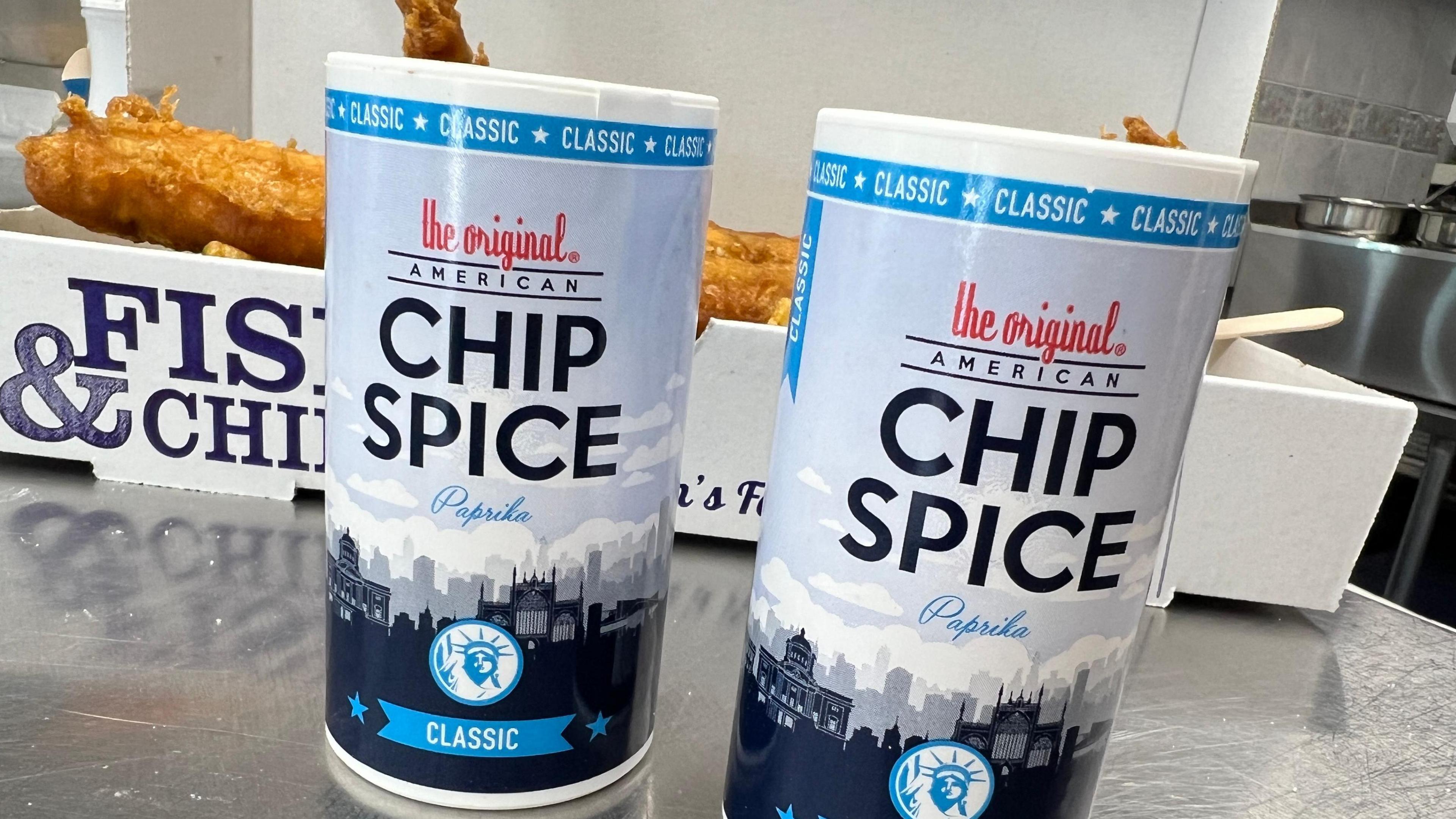 Two pots of American Chip Spice on the chrome-coloured counter of a fish and chip shop, with a box of fried fish and chips in the background. The pots are coloured in shades of blue and feature the outline of the Hull cityscape.