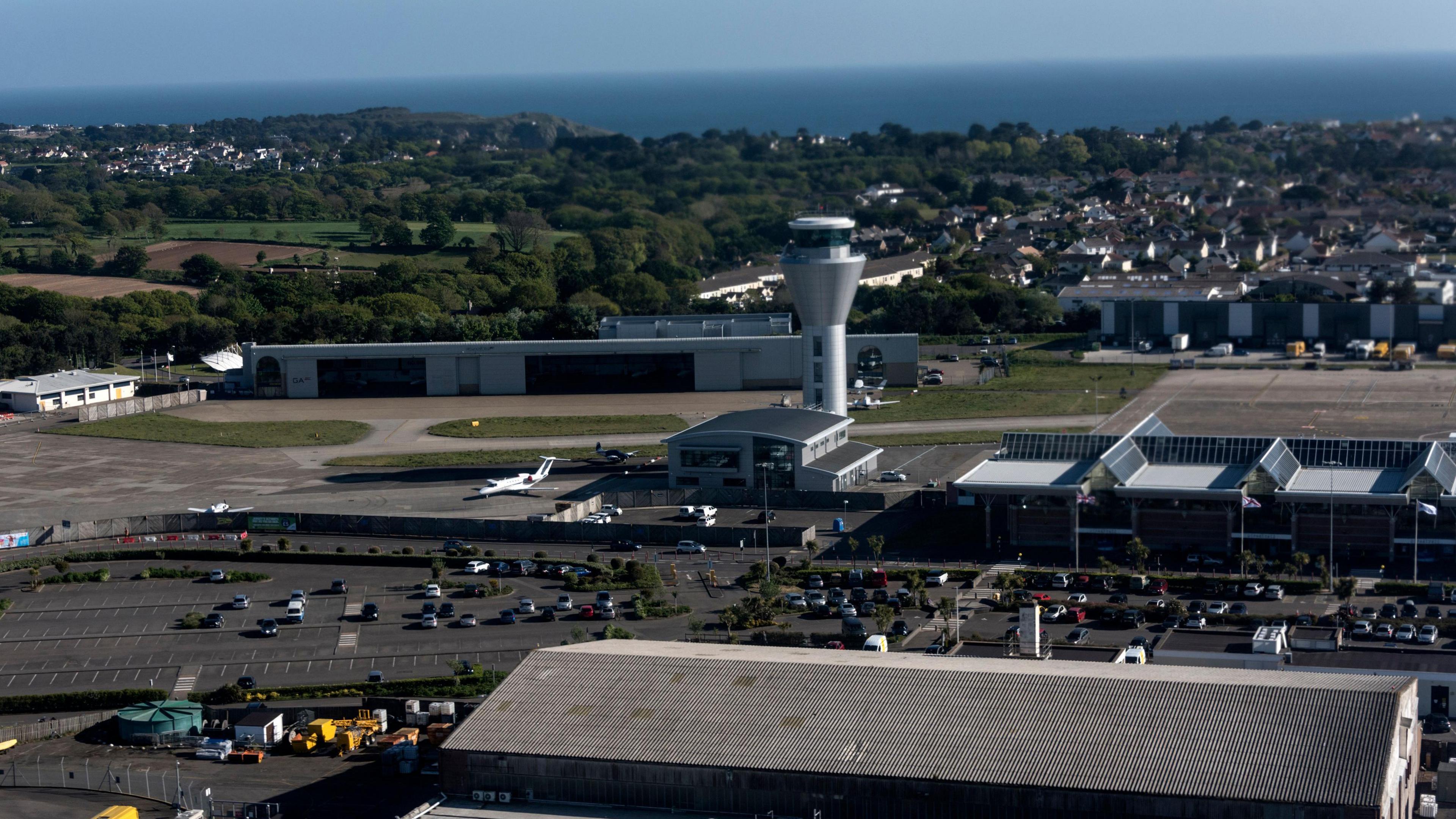 An aerial view of Jersey Airport which shows a large car park to the left with the glass airport building to the right with a triangular glass roof. Behind the airport building is the spherical shaped silver control tower. In the background are houses and trees and behind them is the sea.
