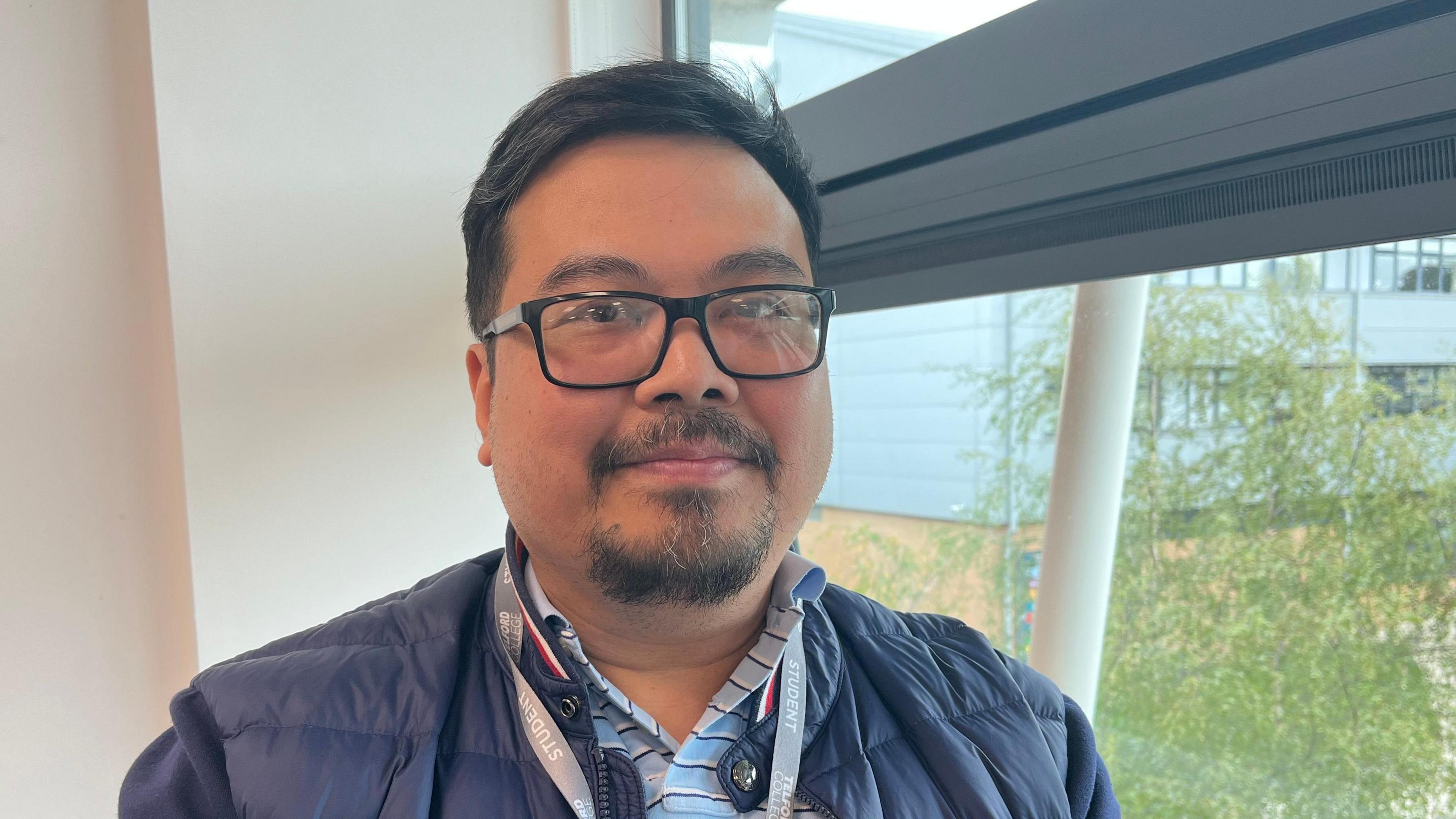 Regino pictured looking into the camera and smiling slightly. He's wearing a blue and white striped collared shirt and a navy blue puffer jacket. He's also got a grey Telford College lanyard on. He's stood by a window, and outside you can see trees and more modern campus buildings.