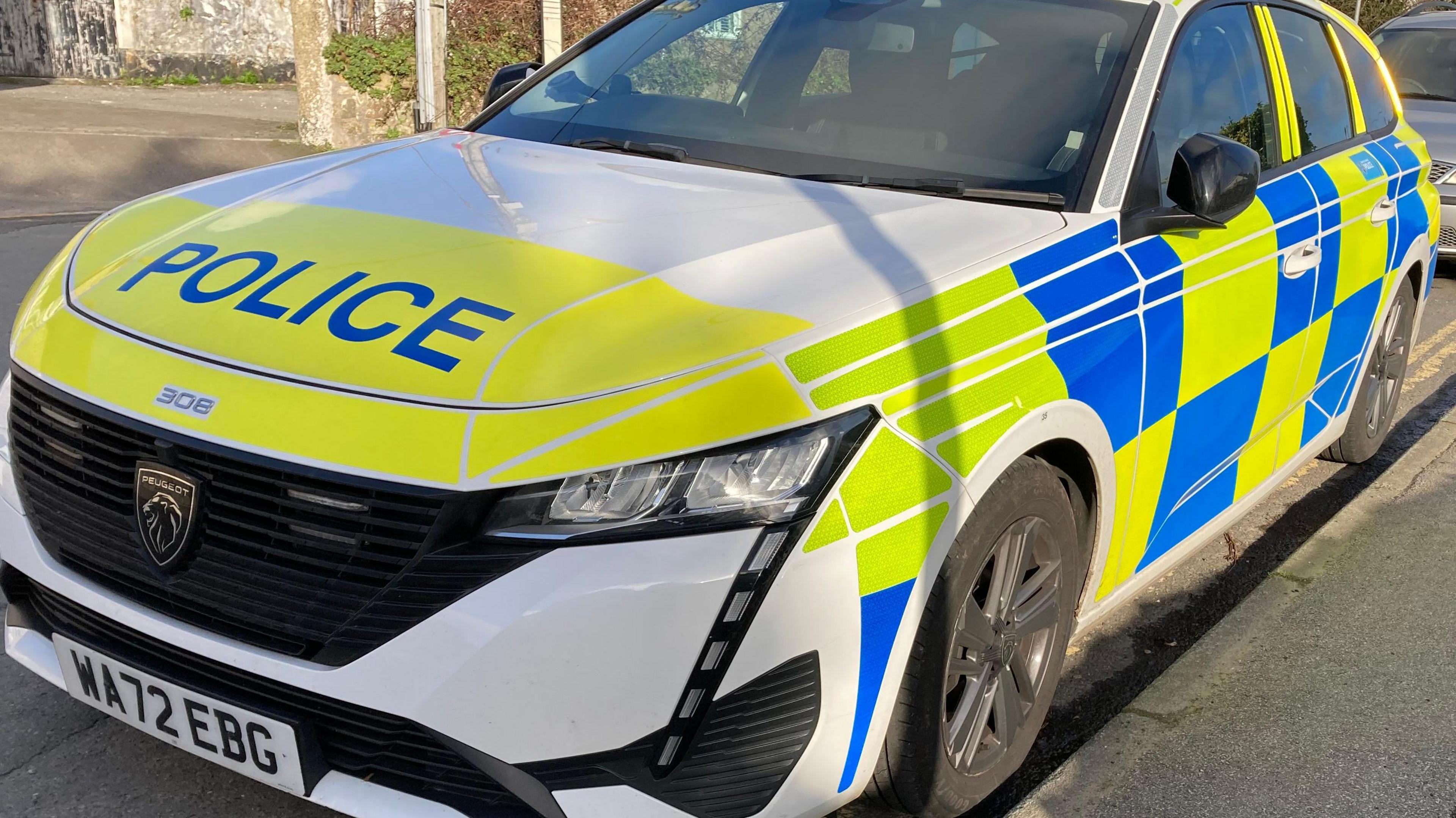 A Devon and Cornwall Police car parked on a residential street. It is white, yellow and blue. The make of the car is Peugeot.