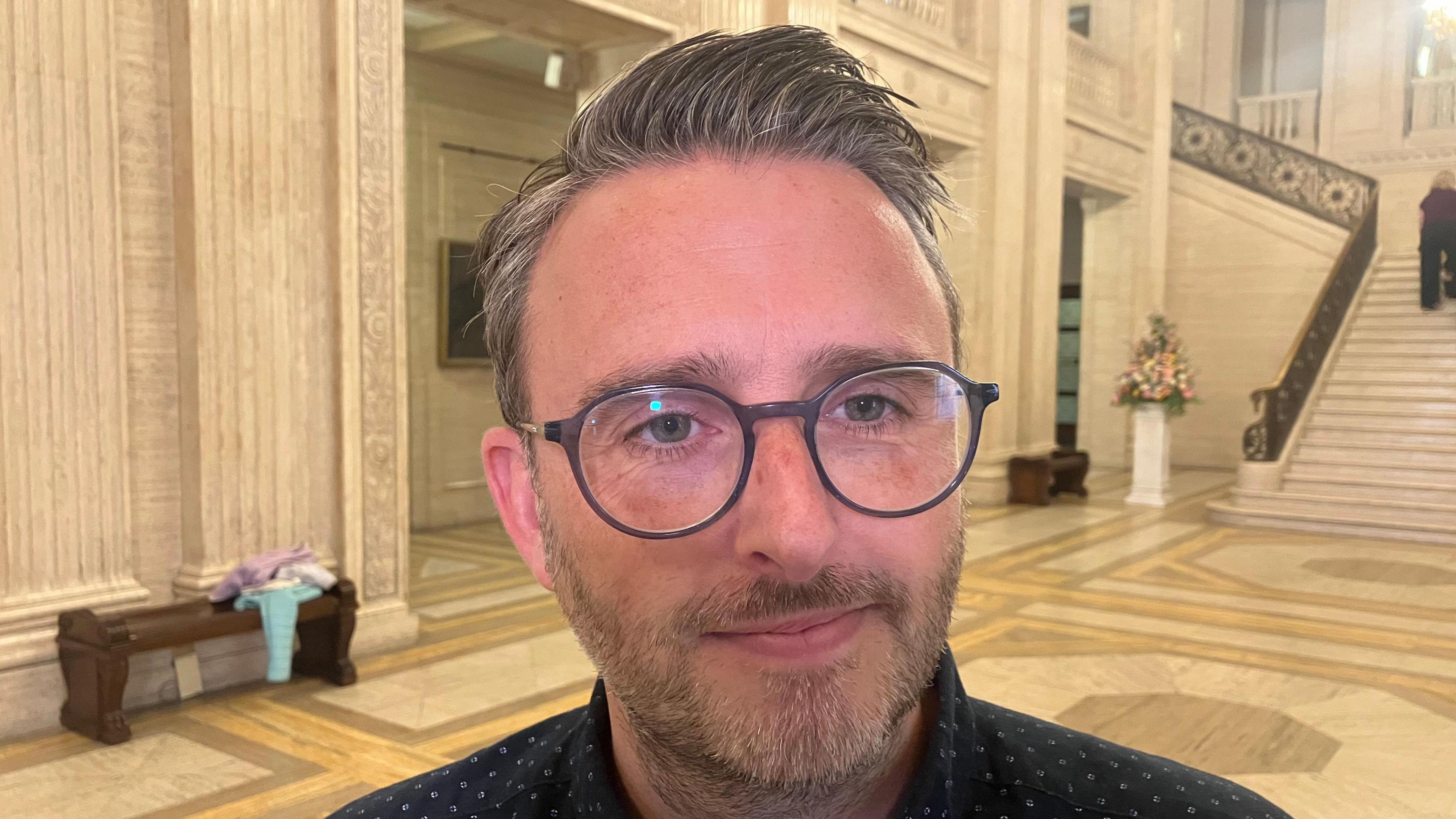 Nick Matheson has a grey beard, grey/whitish hair that is gelled into a spike and grey round glasses. He is wearing a navy shirt and behind him is the yellow white panelled hall and stairs of Stormont.