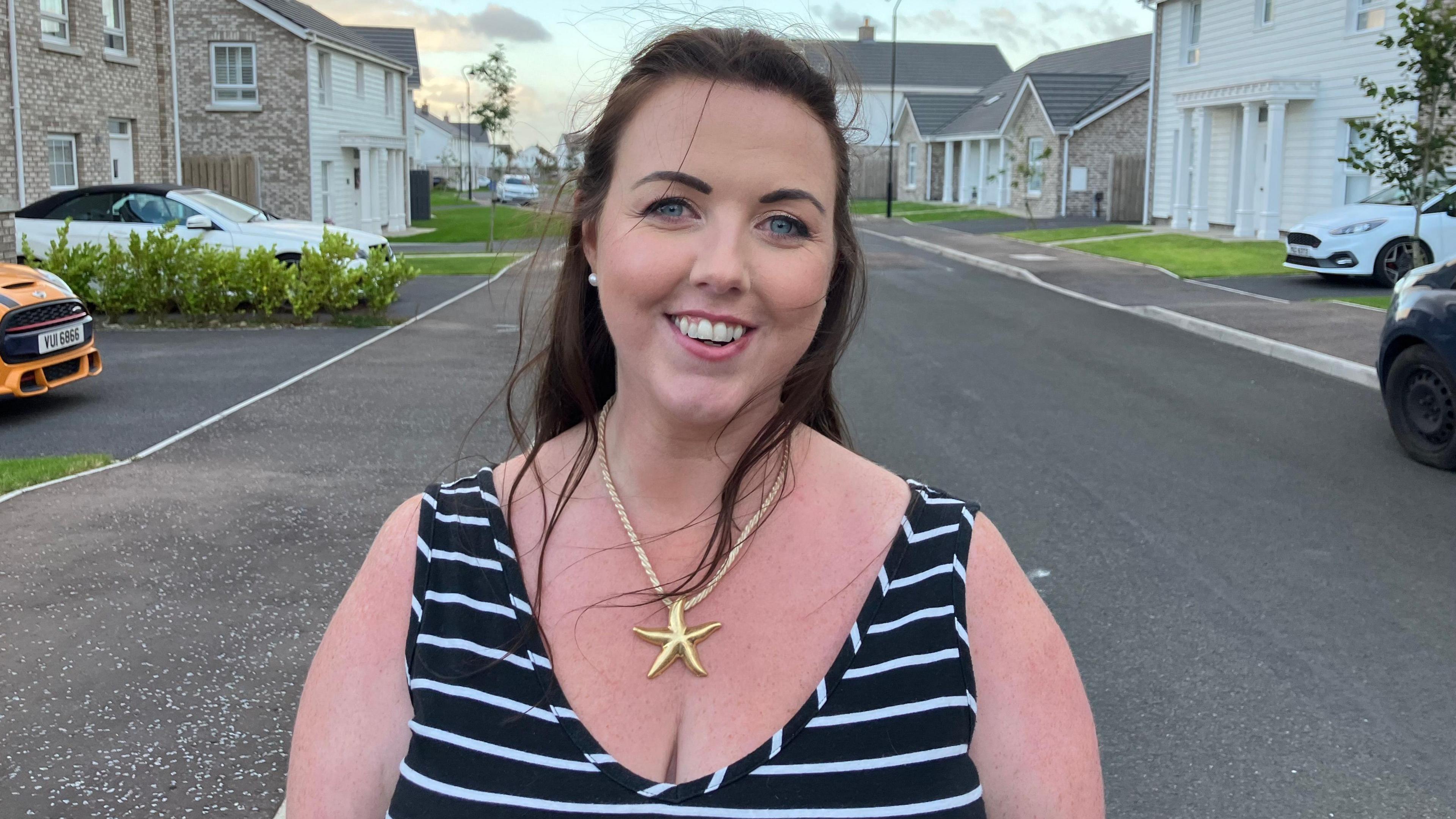 Stephanie O'Fee, wearing a gold necklace with a starfish pendant and a black and white striped strappy top
In housing estate, stood on pavement