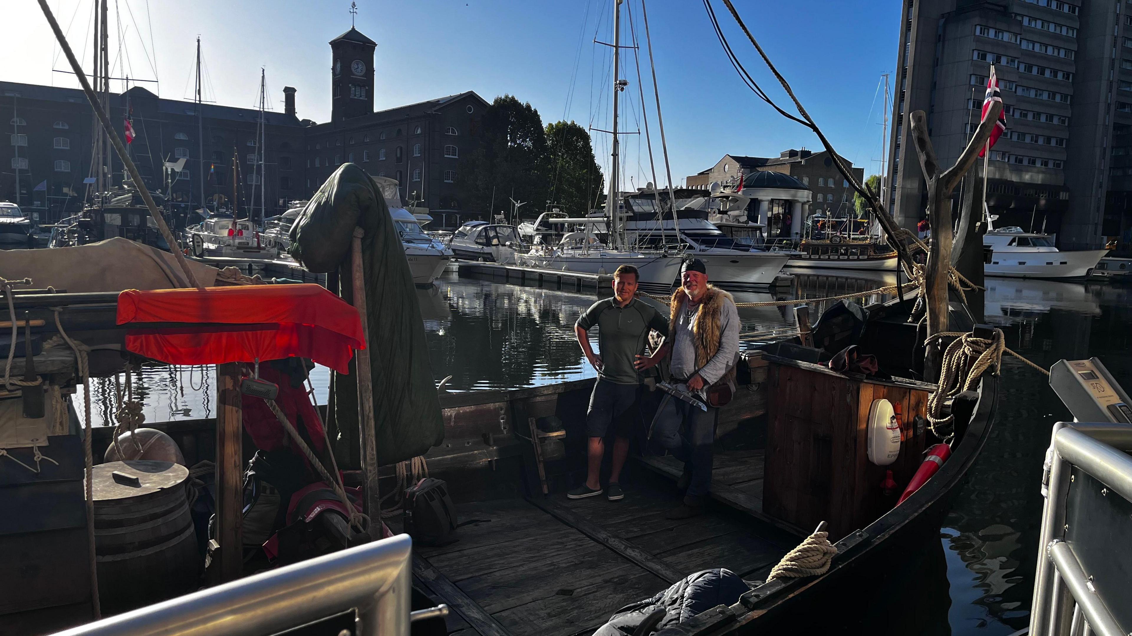 Two men are stood in the distance on board a small wooden ship. The ship is in dock on the water, and the photo is taken from the harbourside beside the boat. Other modern boats are visible in the background.