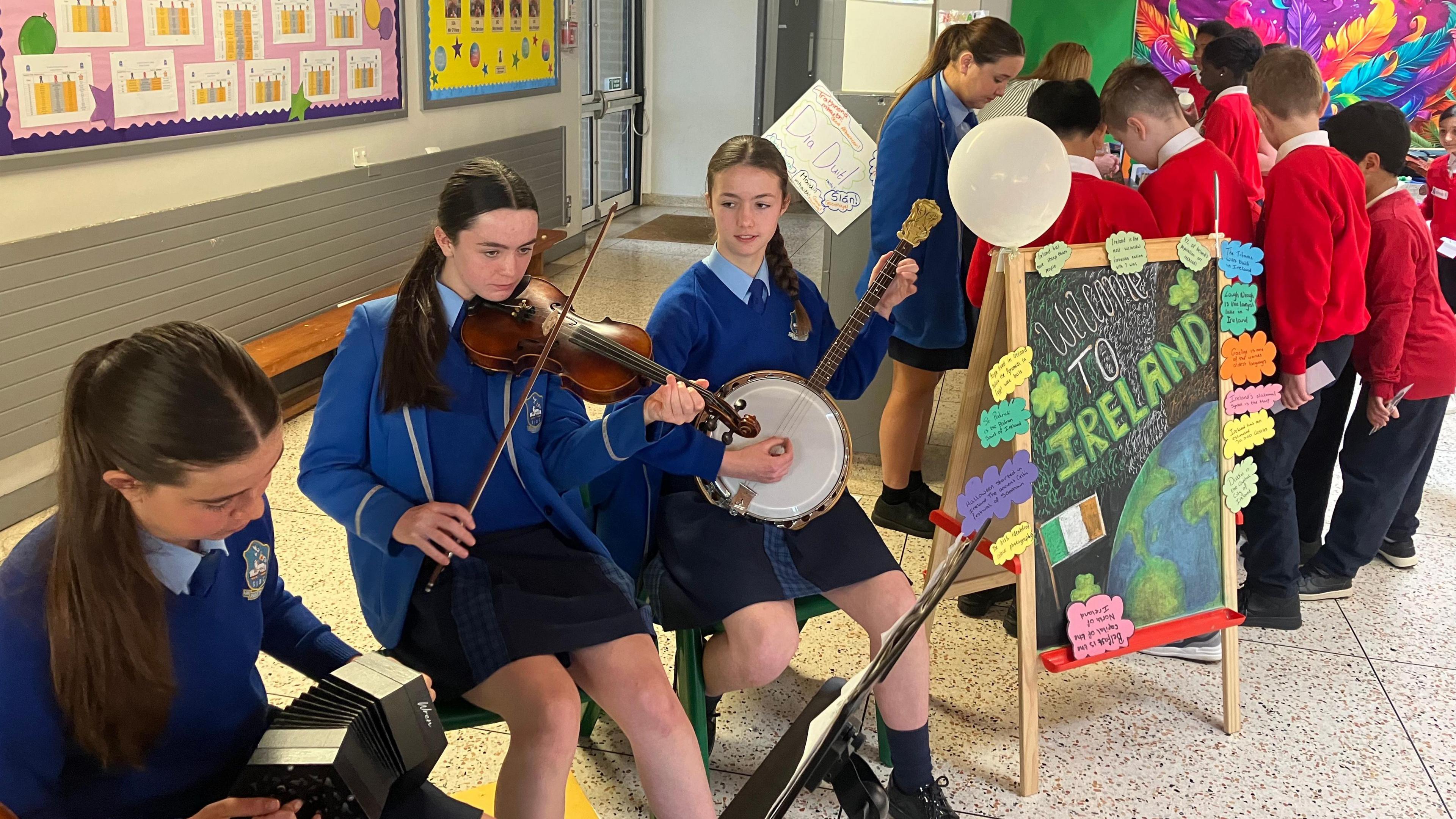 Three girls playing instruments. One on the left is playing the accordion, one in the middle is playing the violin and the one on the right is playing a banjo. All have long dark brown hair tied back and wearing blue school uniforms. A sign saying 'Welcome To Ireland' is beside them.