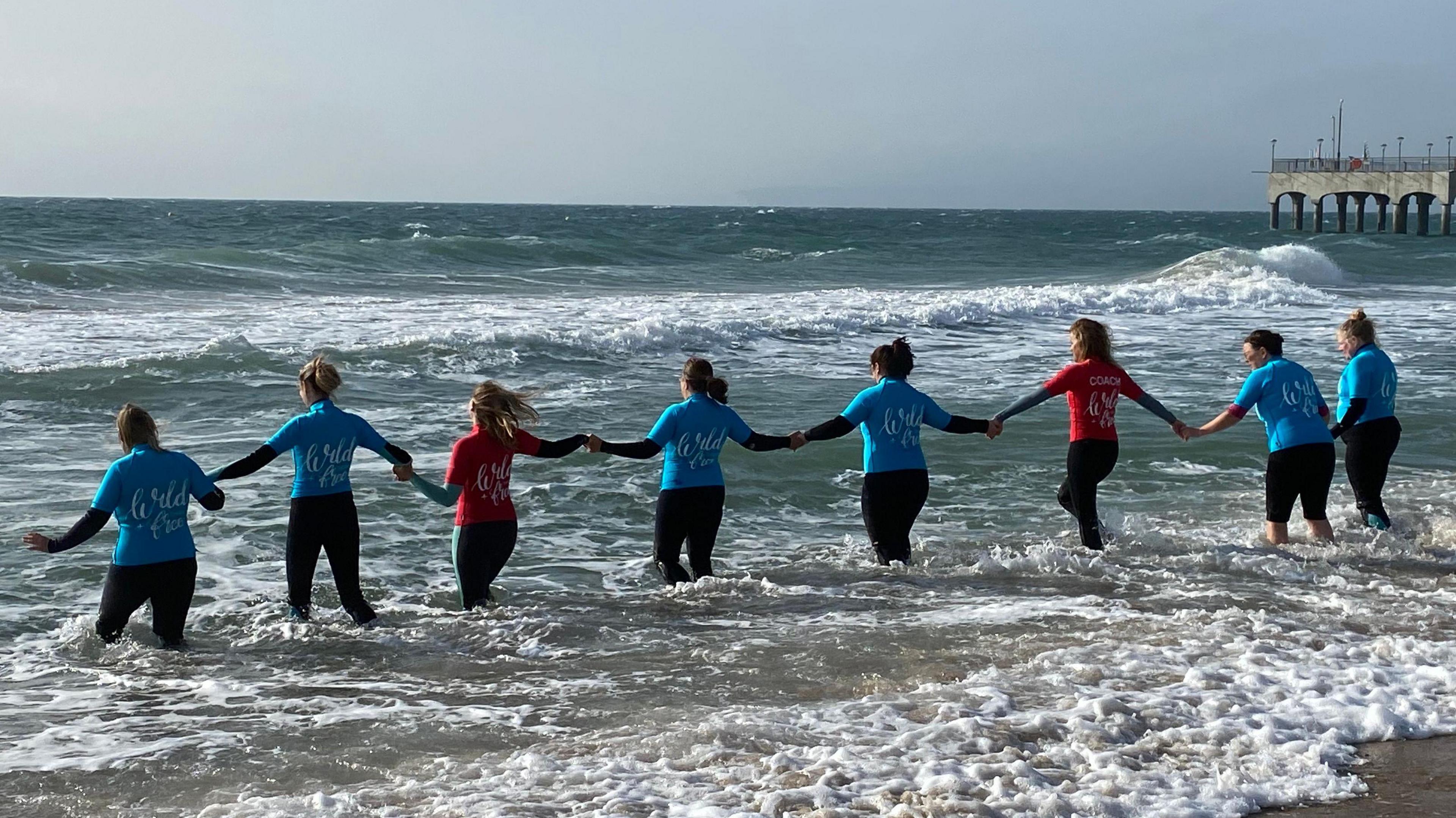 A group of people stood knee high in the sea facing backwards, with a pier in the background