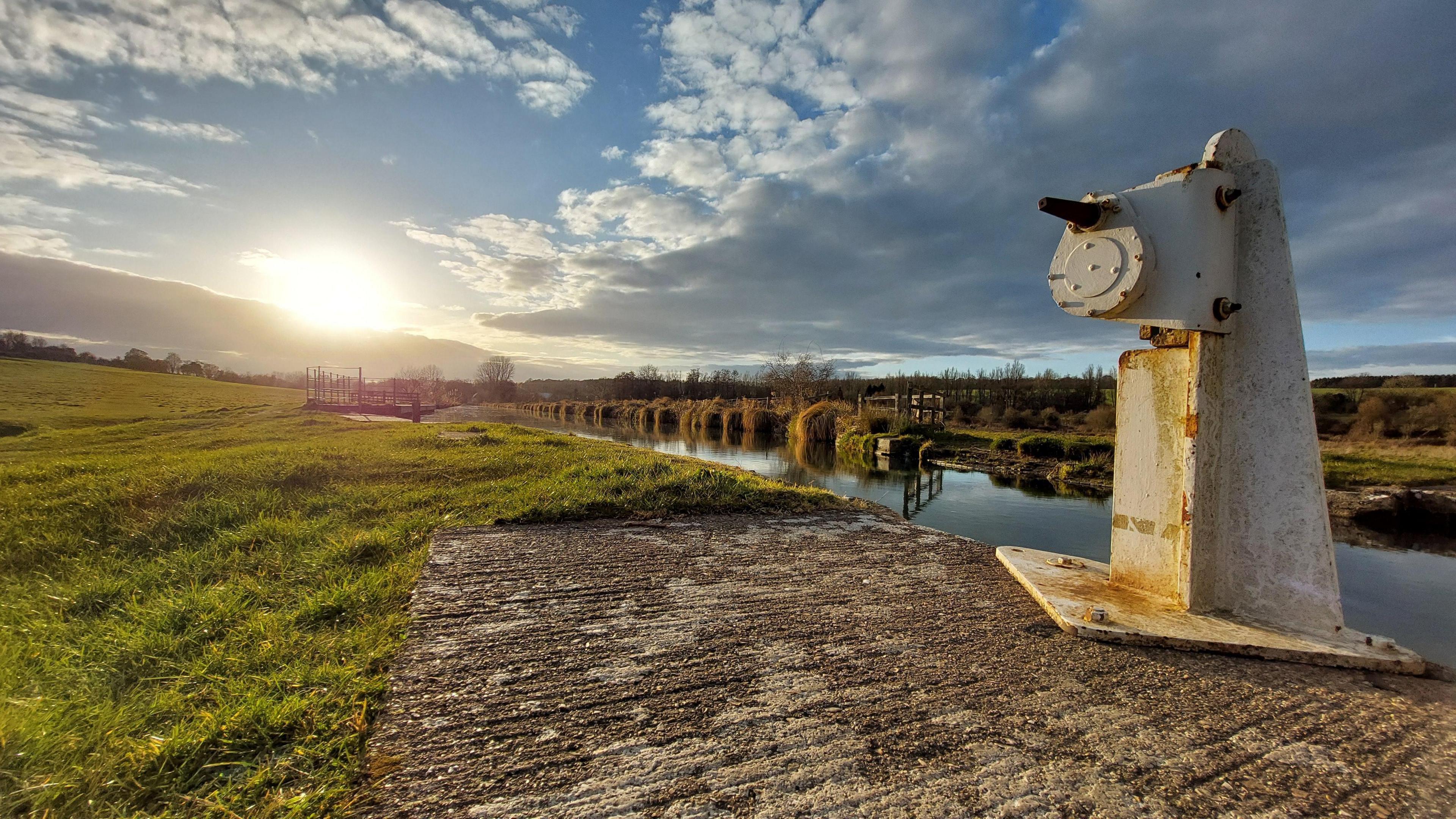 The grass bank of the canal is in the foreground with a white mooring post. The sun is setting over the horizon with a blue sky that has some cloud cover. 