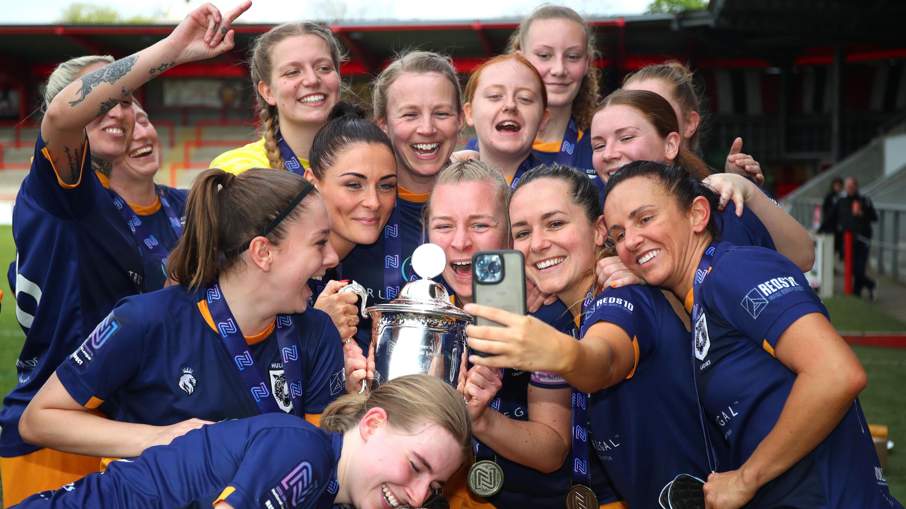 A group of 14 women wearing navy blue football shirts and amber shorts smile and hug as they pose for a selfie with a silver trophy. The picture shows women smiling at phone taking a selfie. They have medals around their necks.