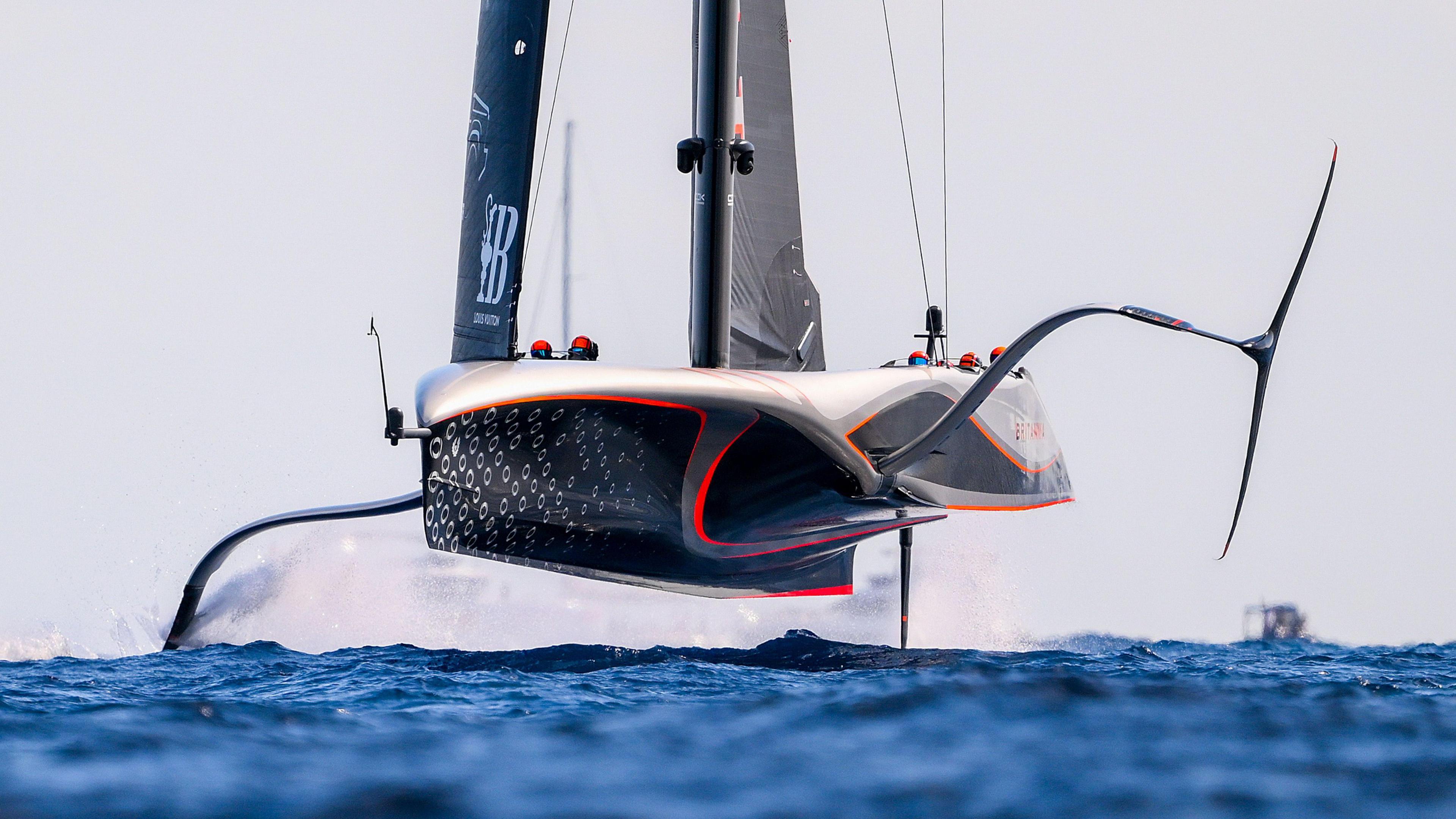 Ineos Britannia America's Cup boat lifts out of the water with one of his hydrofoil wings visible. The boats hull is silver and black with red markings and the sea is a bright blue