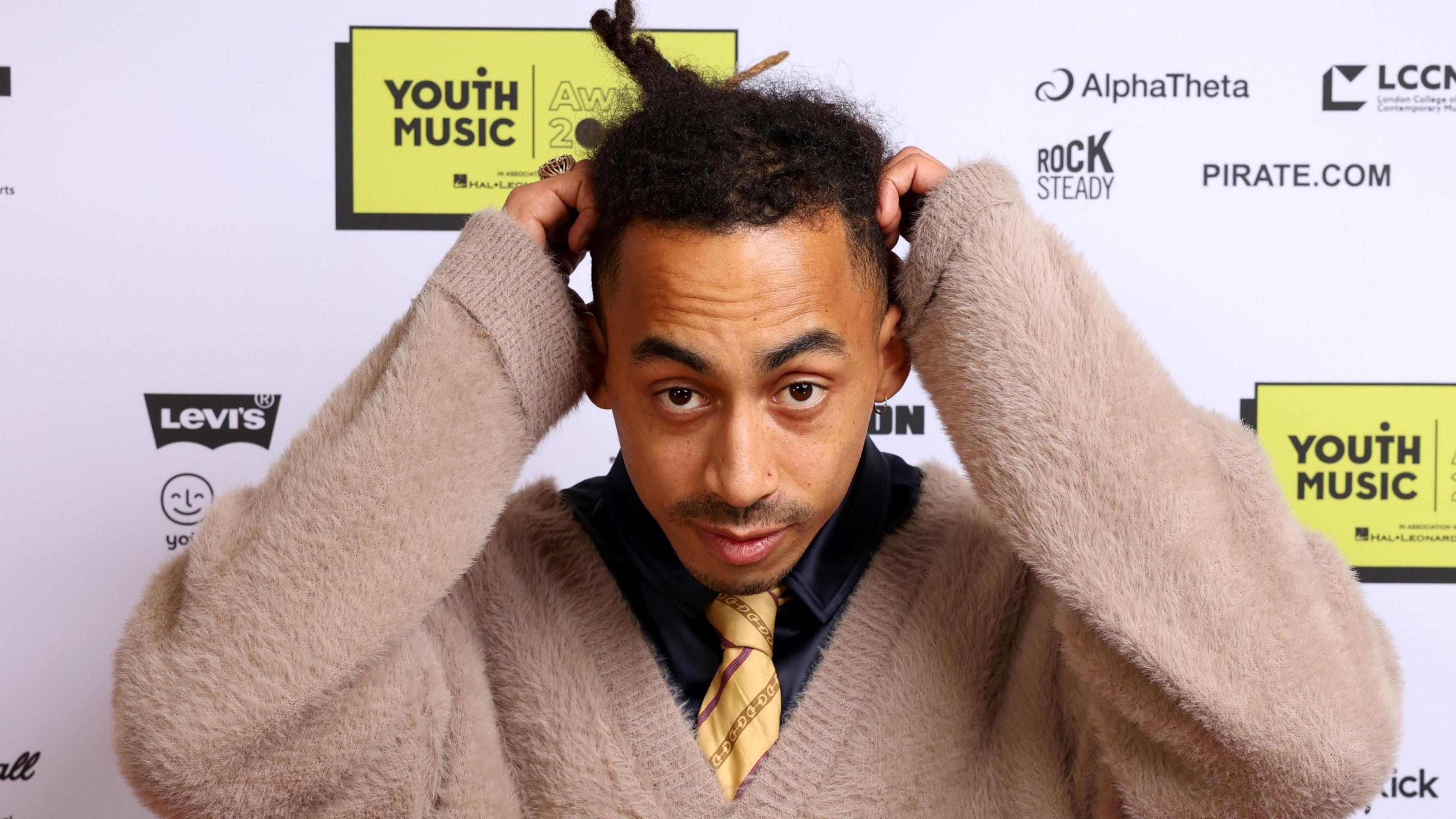 Jordan Stephens at the Youth Music Awards. Jordan wears a fluffy beige cardigan over a navy shirt and yellow striped tie. He holds his hands to his head as he looks at the camera.