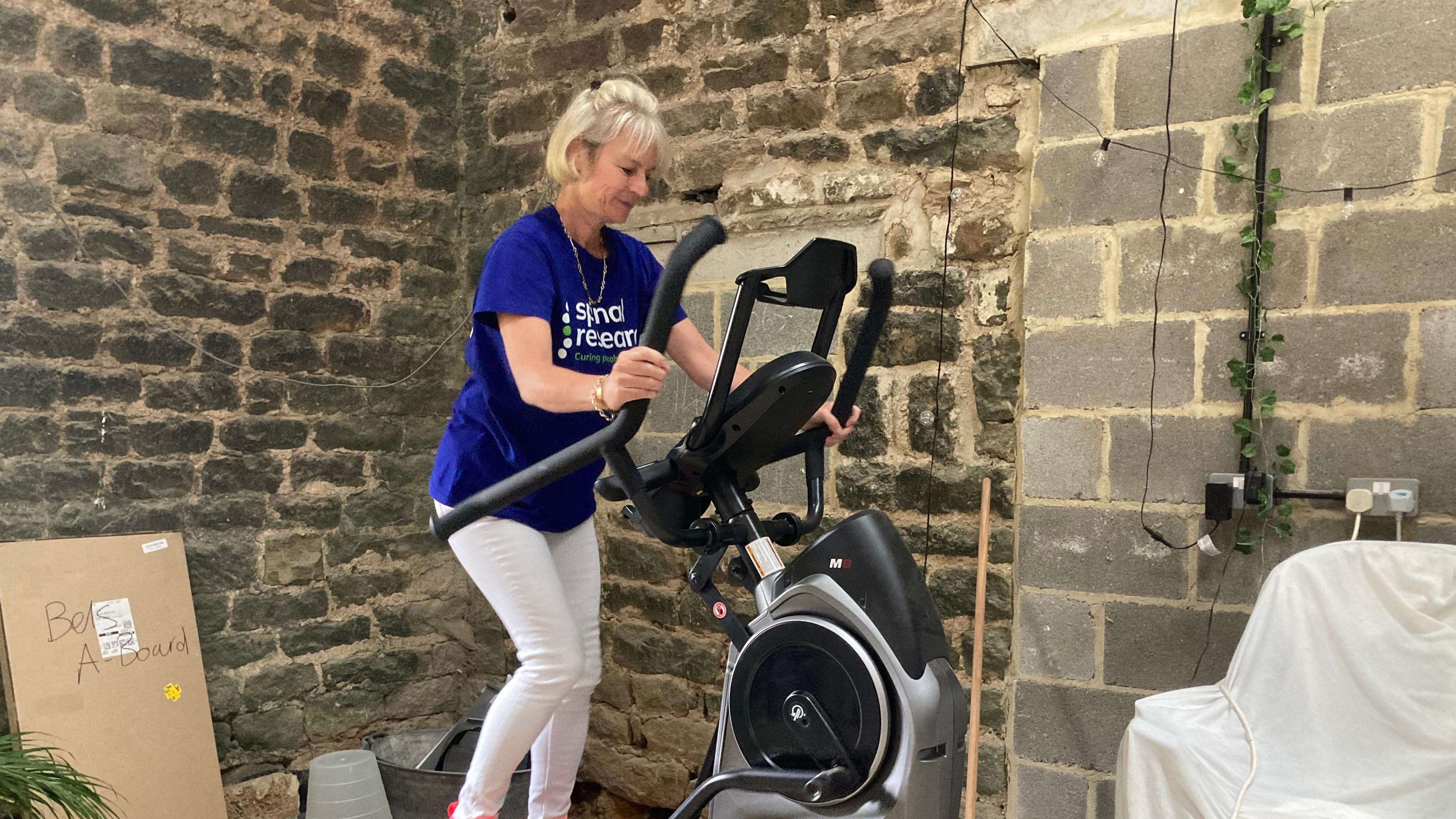 A woman in her 50s on a cross trainer. She has blonde hair and is wearing a blue T-shirt and white trousers. She is next to an old brick wall.