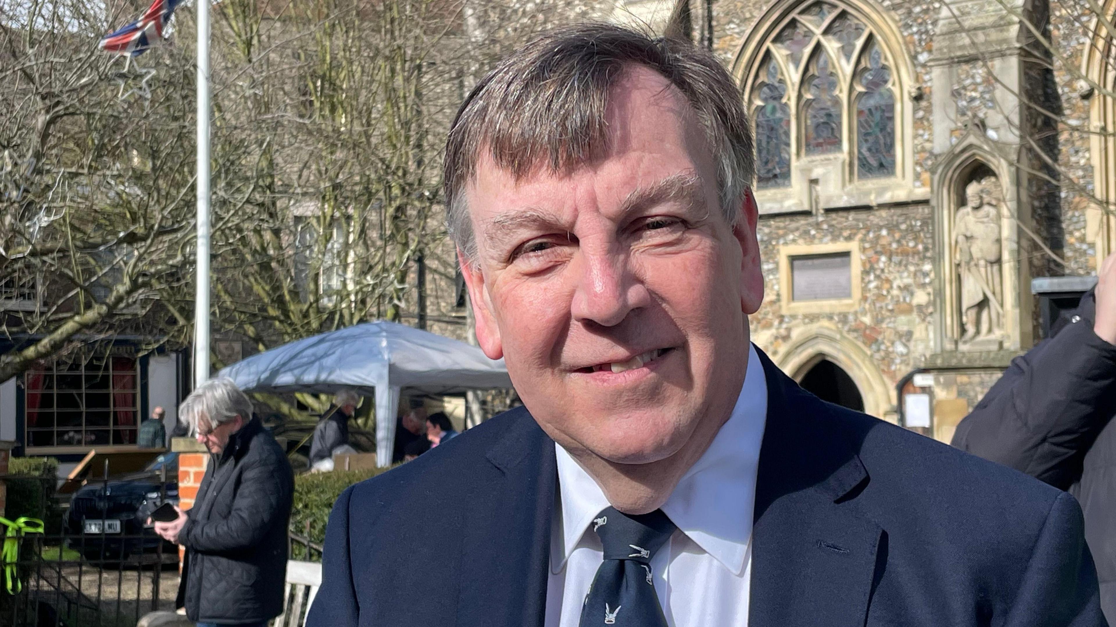 John Whittingdale who is wearing a black suit with a white shirt and black tie. He is smiling at the camera.