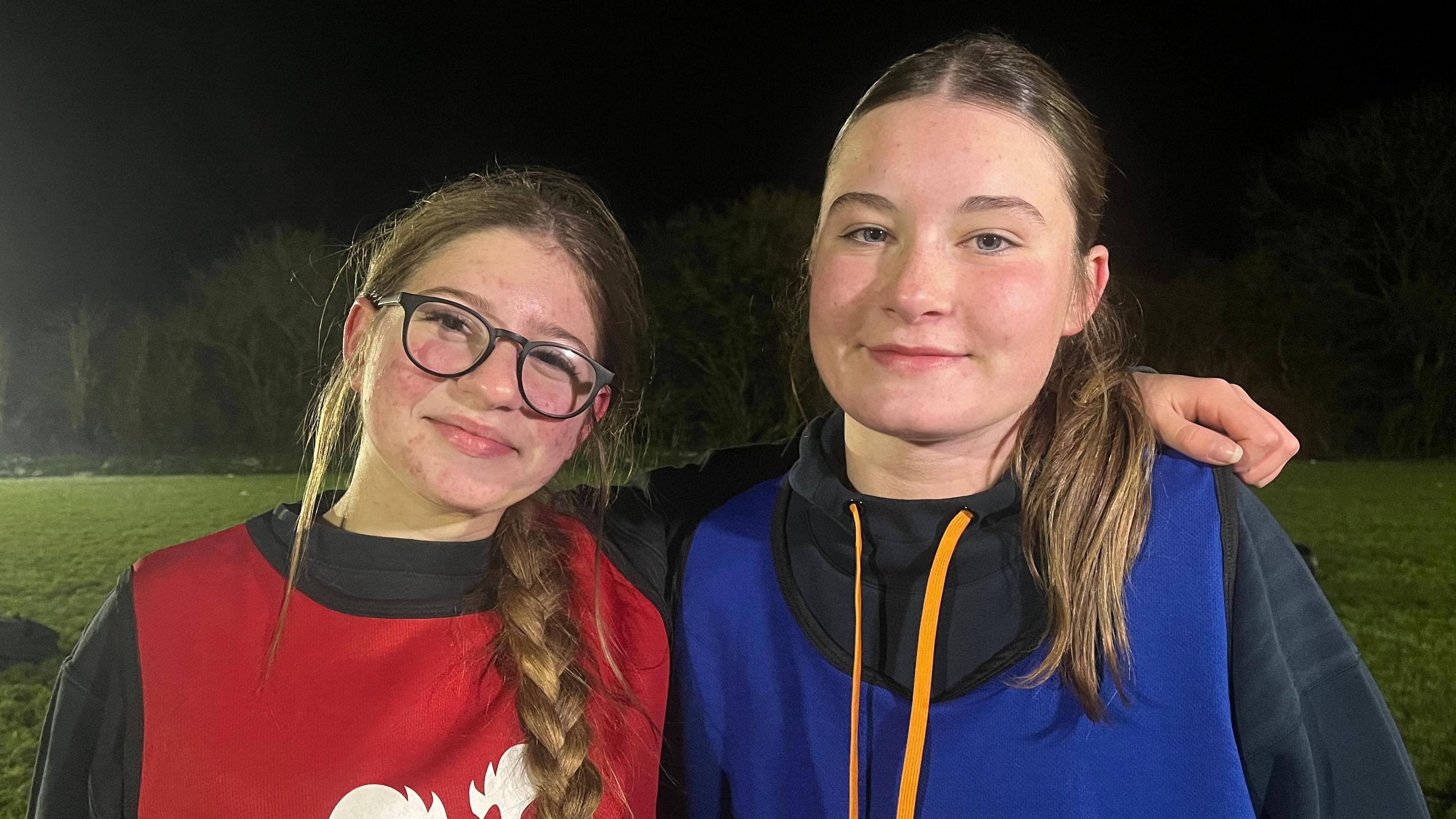 Esme and Megan stood side by side, smiling slightly and looking into the camera. They're both wearing black sweatshirts - Esme, on the left, is wearing a red tabard, and Megan, on the right, is wearing a blue one. They're stood on a flood-lit rugby pitch, which has green grass, a dark night sky, and is bordered by trees.