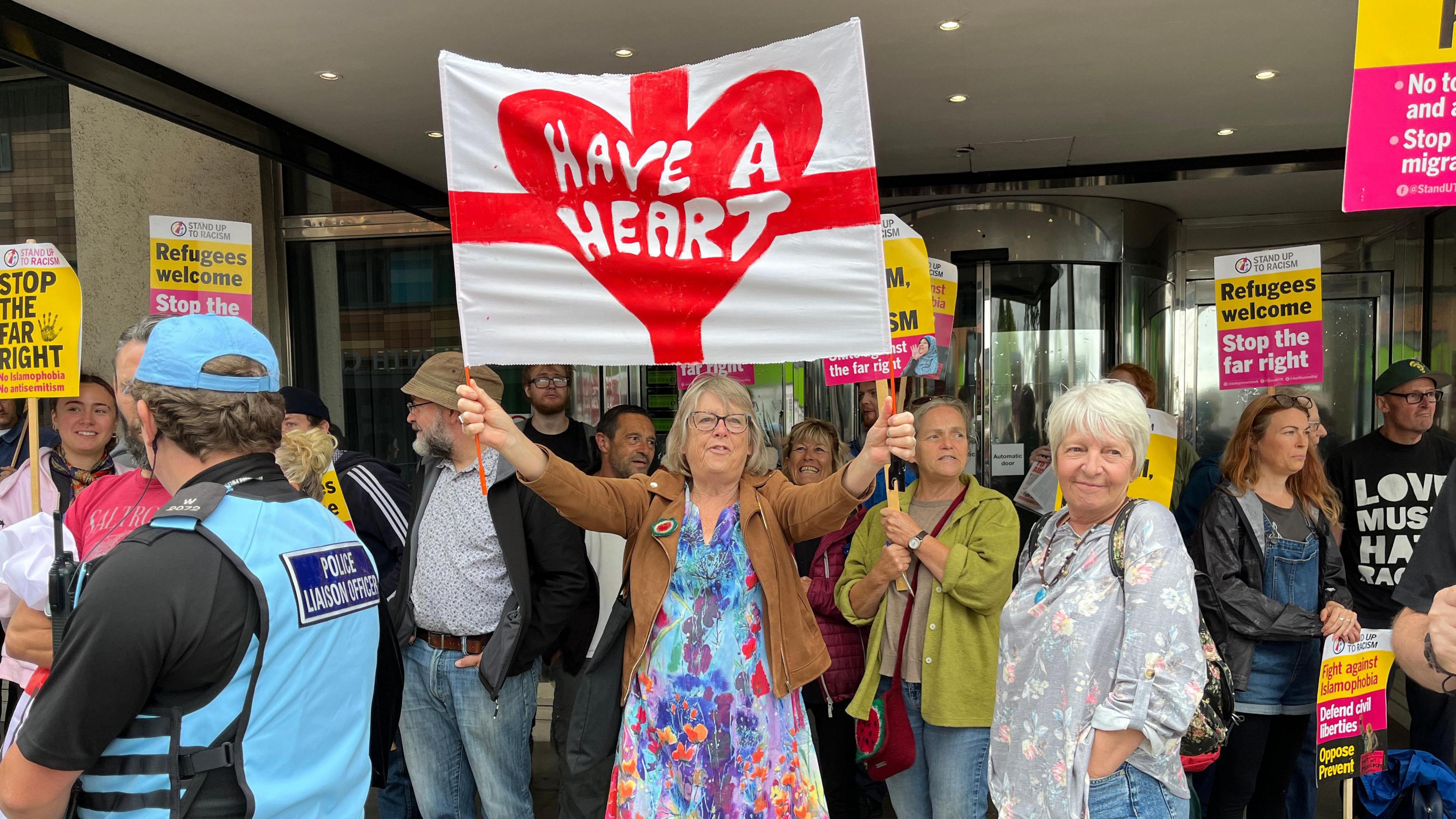 A group of people stand outside a set of revolving doors. A woman in a brightly coloured dress holds an England flag with the words "Have a heart" on it. Other people hold placards saying "Refugees welcome" and "Stop the right".