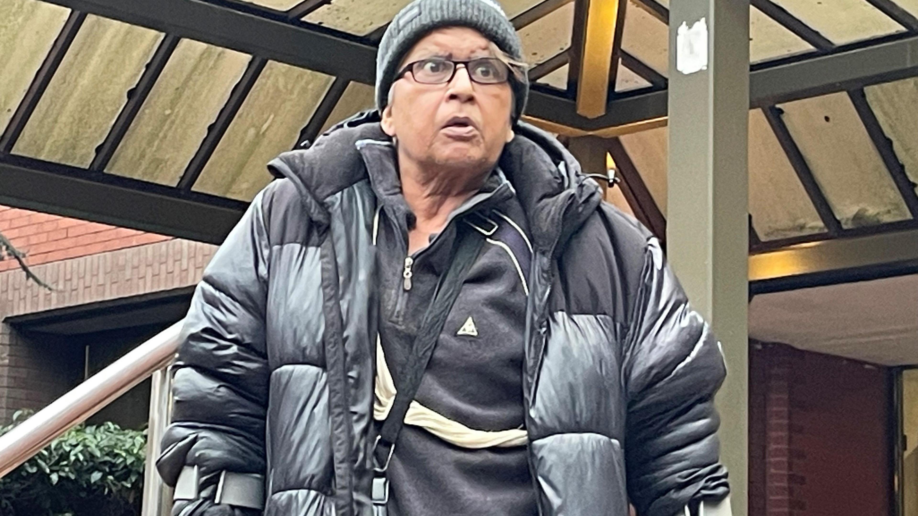 Farhat Ajaz, 62, leaving Birmingham Crown Court. He is wearing a black puffer jacket and woolly hat and is wearing glasses and walking with crutches.