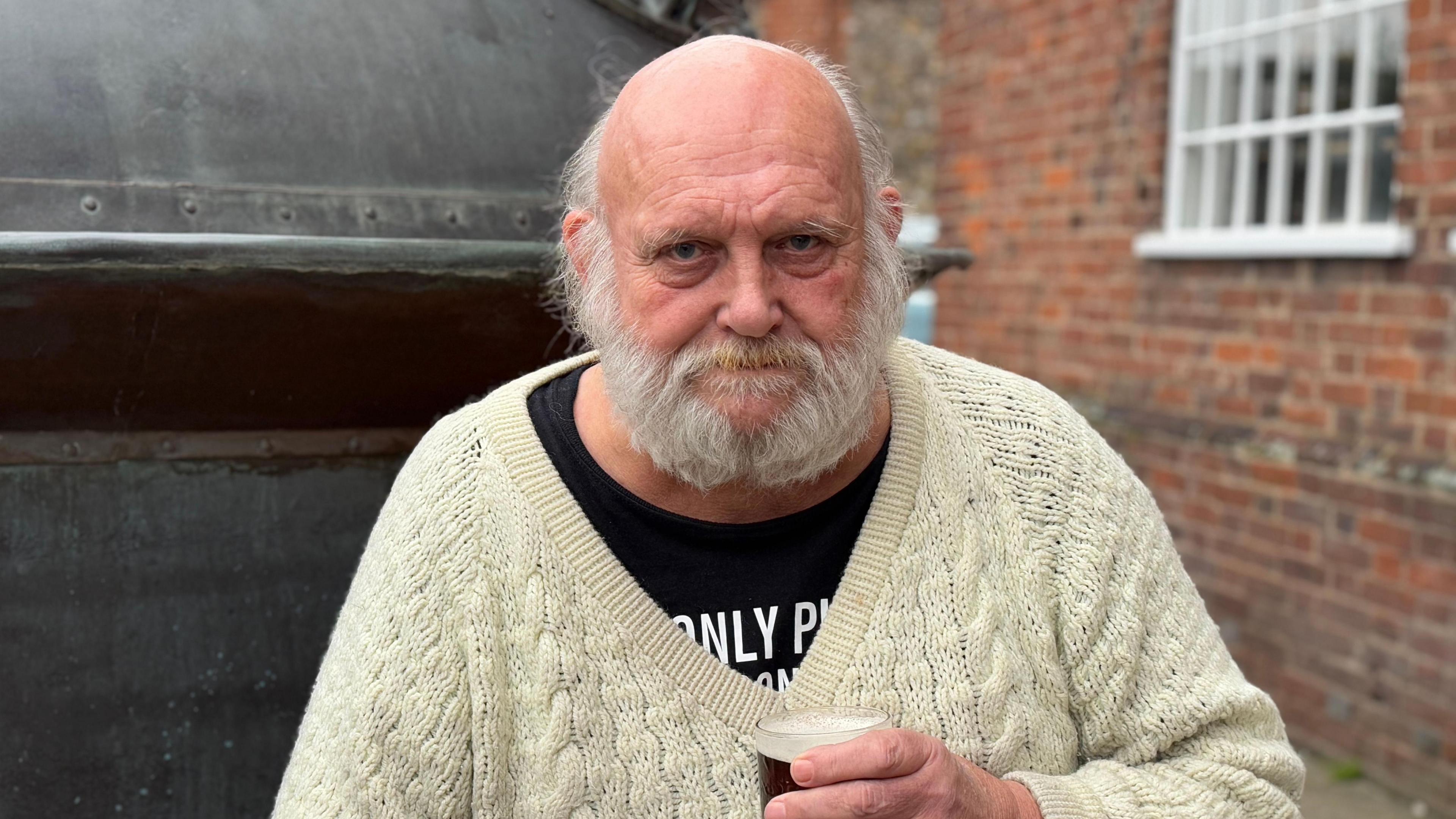 An older man with a grey beard wearing a cream cardigan and holding a pint of beer