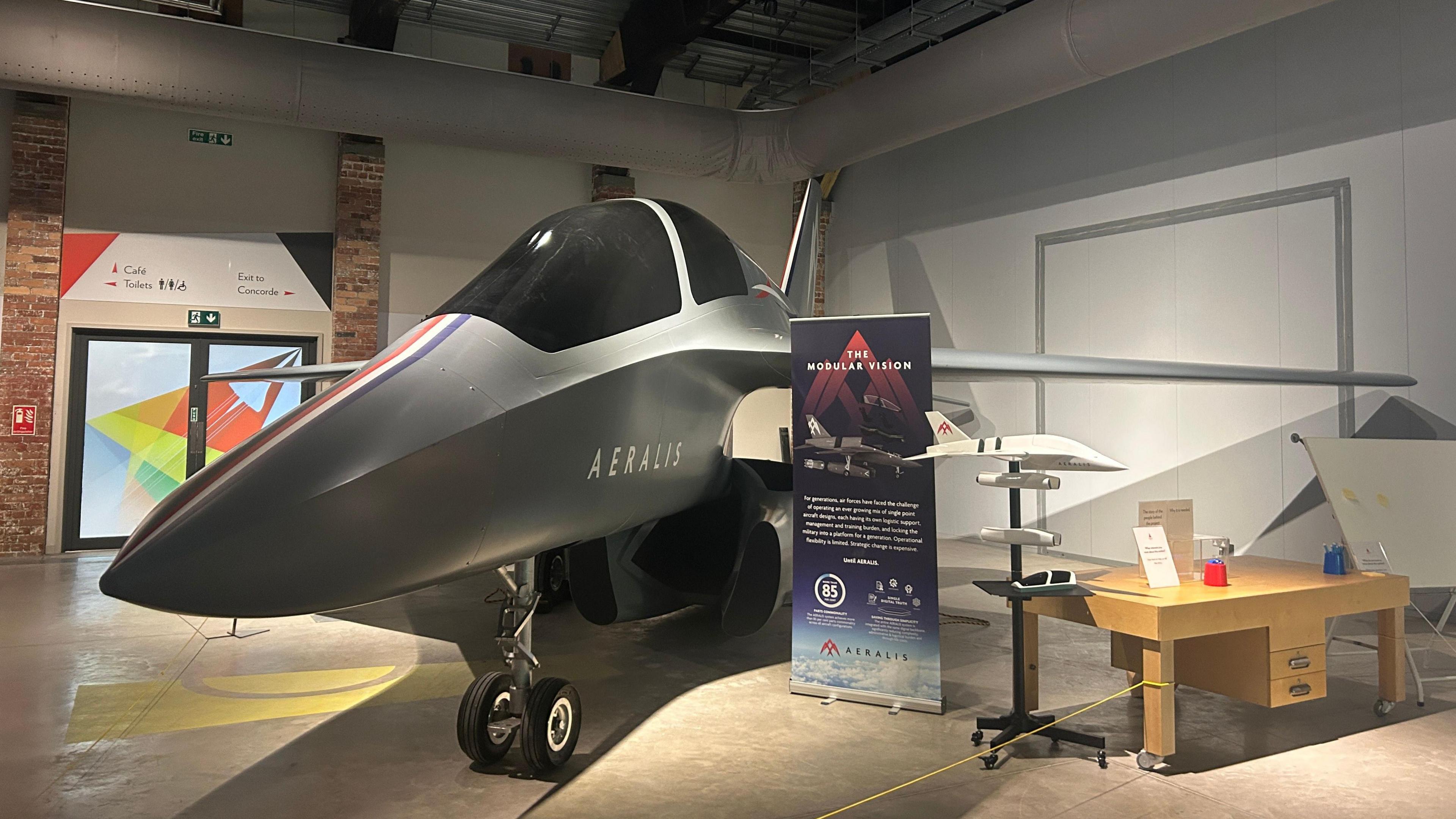 A modern jet-style aircraft is inside the Aerospace Bristol aviation attraction. It is flanked by a board explaining what it is, and a scale model