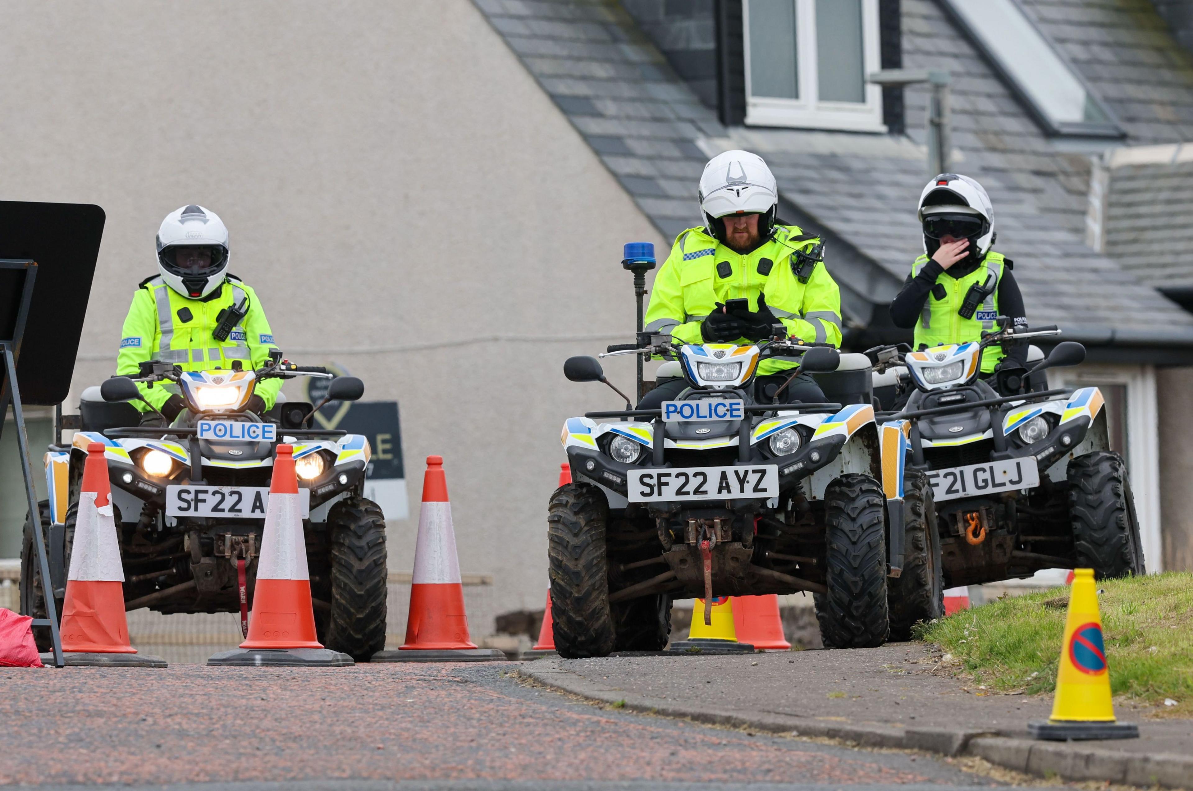 Three police officers on quad bikes with cones near them. They are wearing white helmets and hi-vis jackets