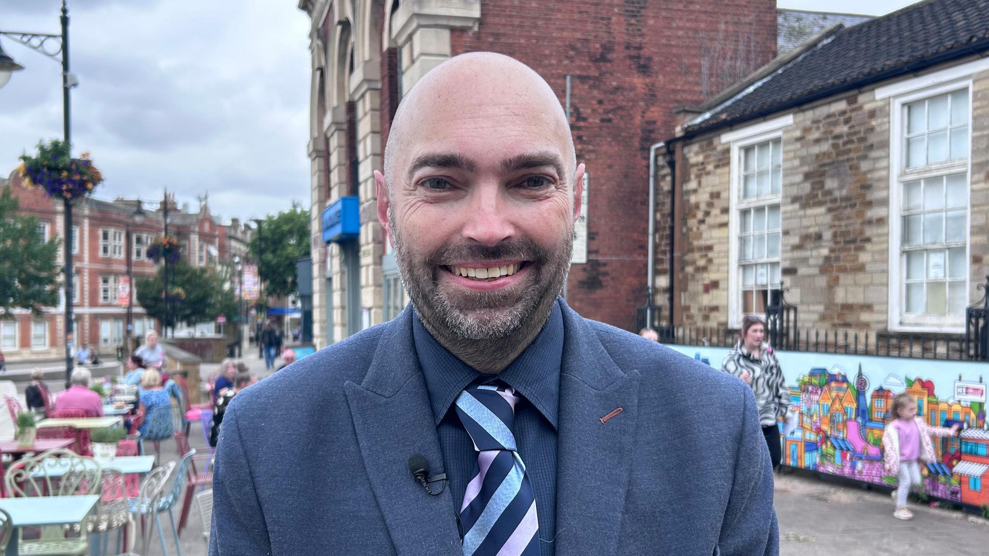 Lee Watkiss, the leader of Kettering Town Council stands in the town square. He is in a navy blue jacket and shirt with a patterned light blue, dark blue and pink tie. He is bald and has a short beard with dark hair.