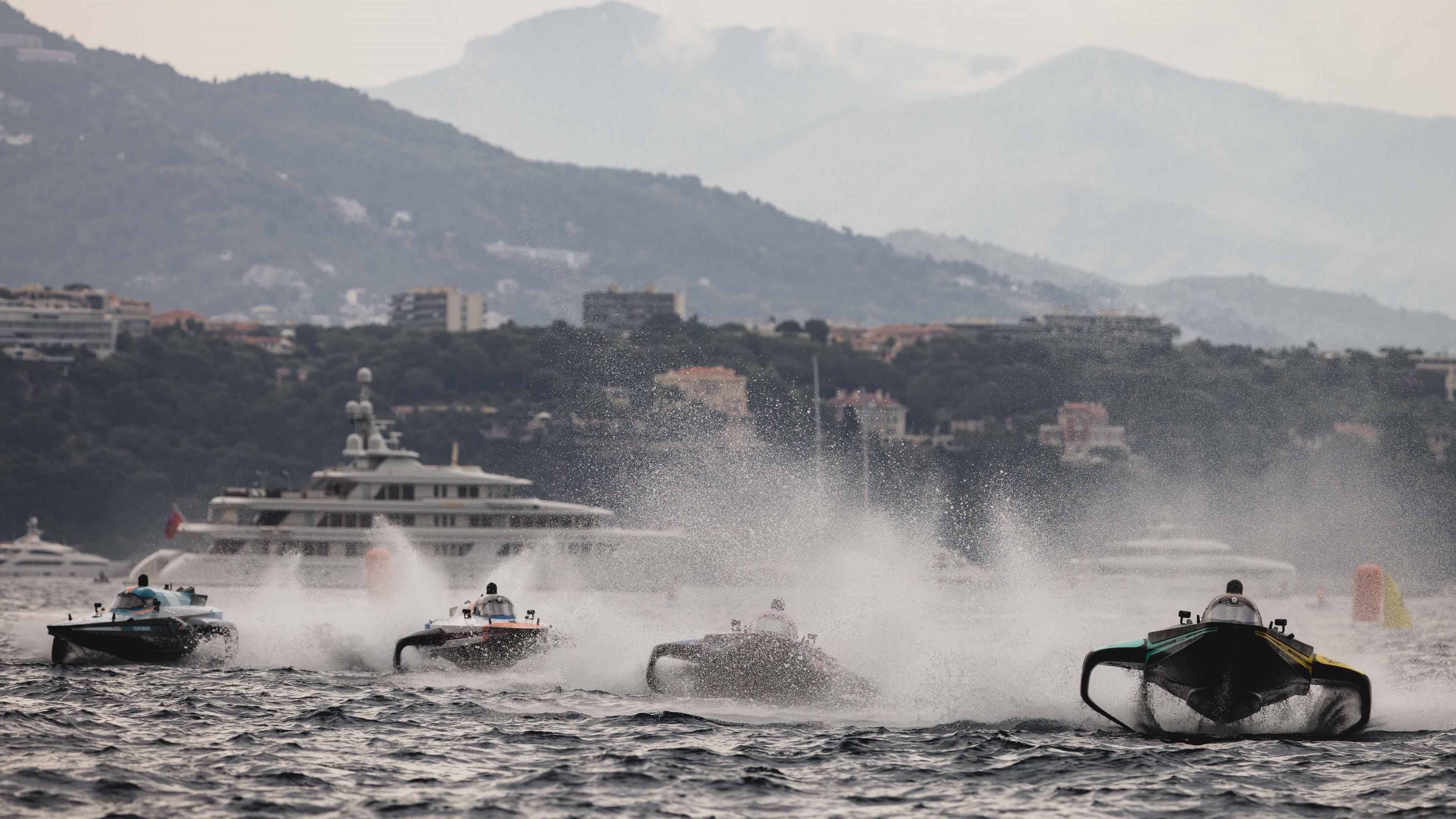 Four twin-hulled powerboats are partially obscured as they throw spray into the air while racing on water. In the background a large yacht can be seen in front of buildings on a green shore line and three hills are seen in the distance