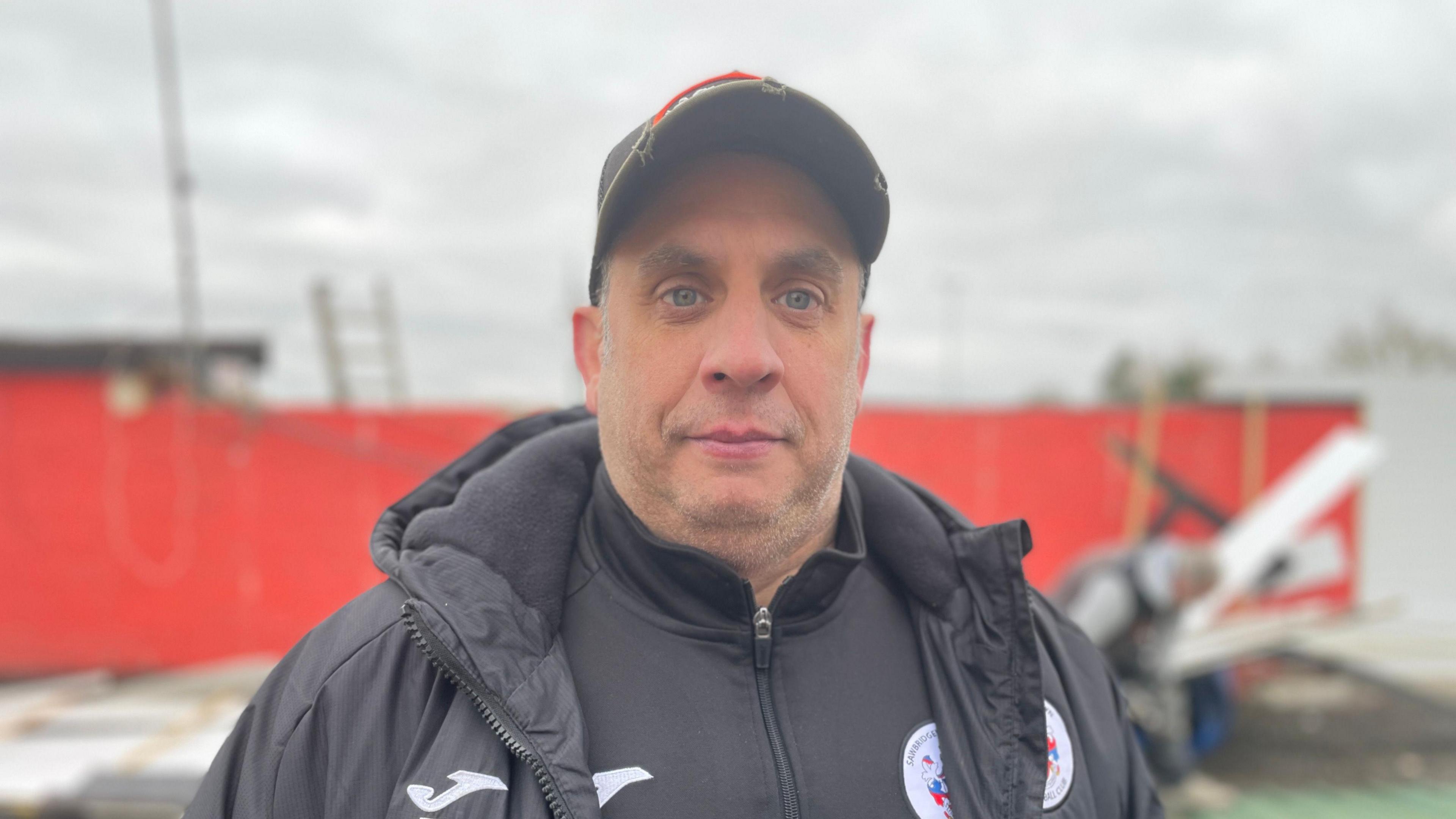 Ross Livermore is wearing a black puffer jacket and a black quarter-zip, both bearing the club crest. He is also wearing a cap. Behind him is a red wall, a ladder and men working.
