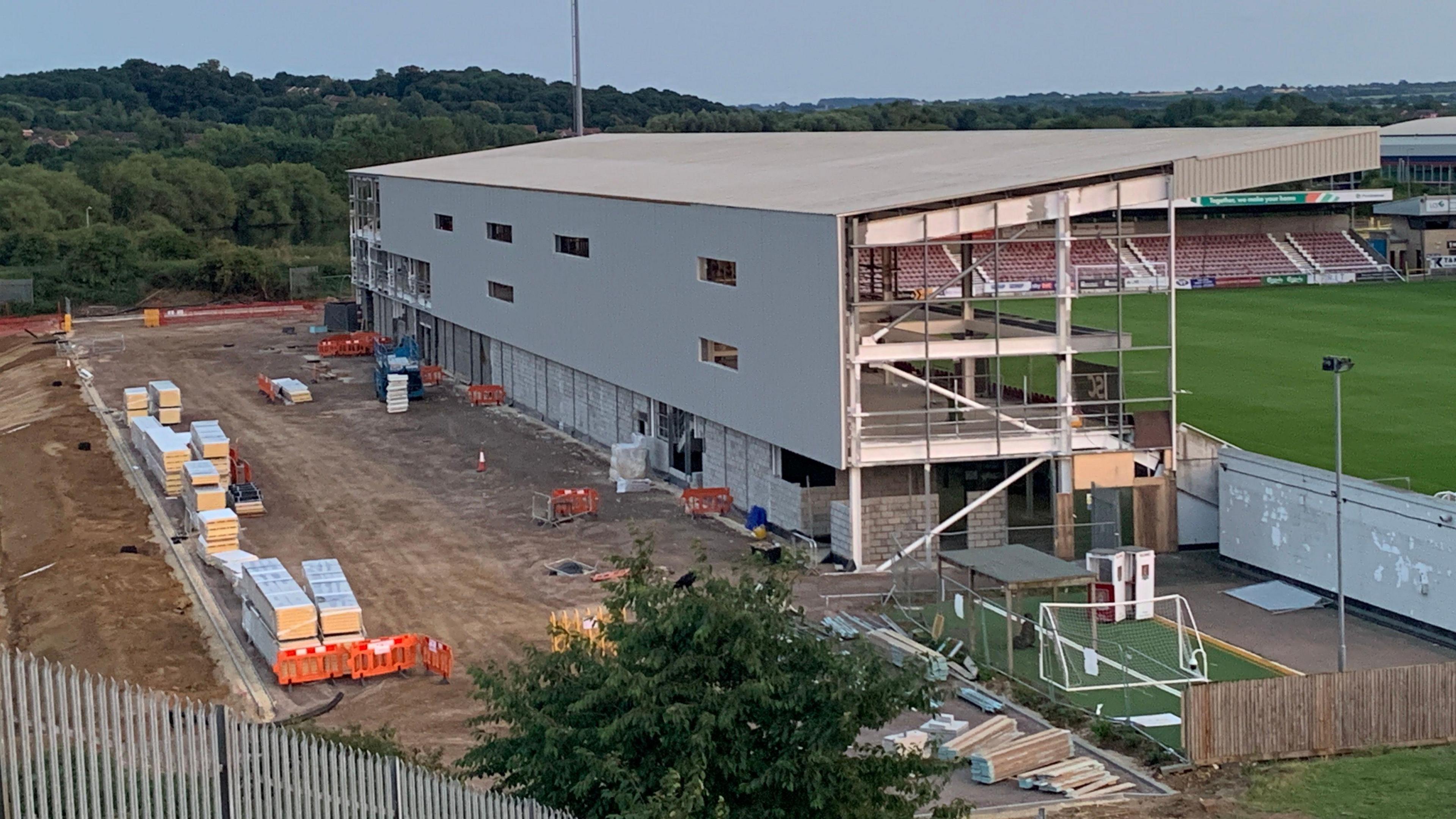 A white stand with scaffolding looking out on to a football pitch. Behind the stand are piles of building materials. 