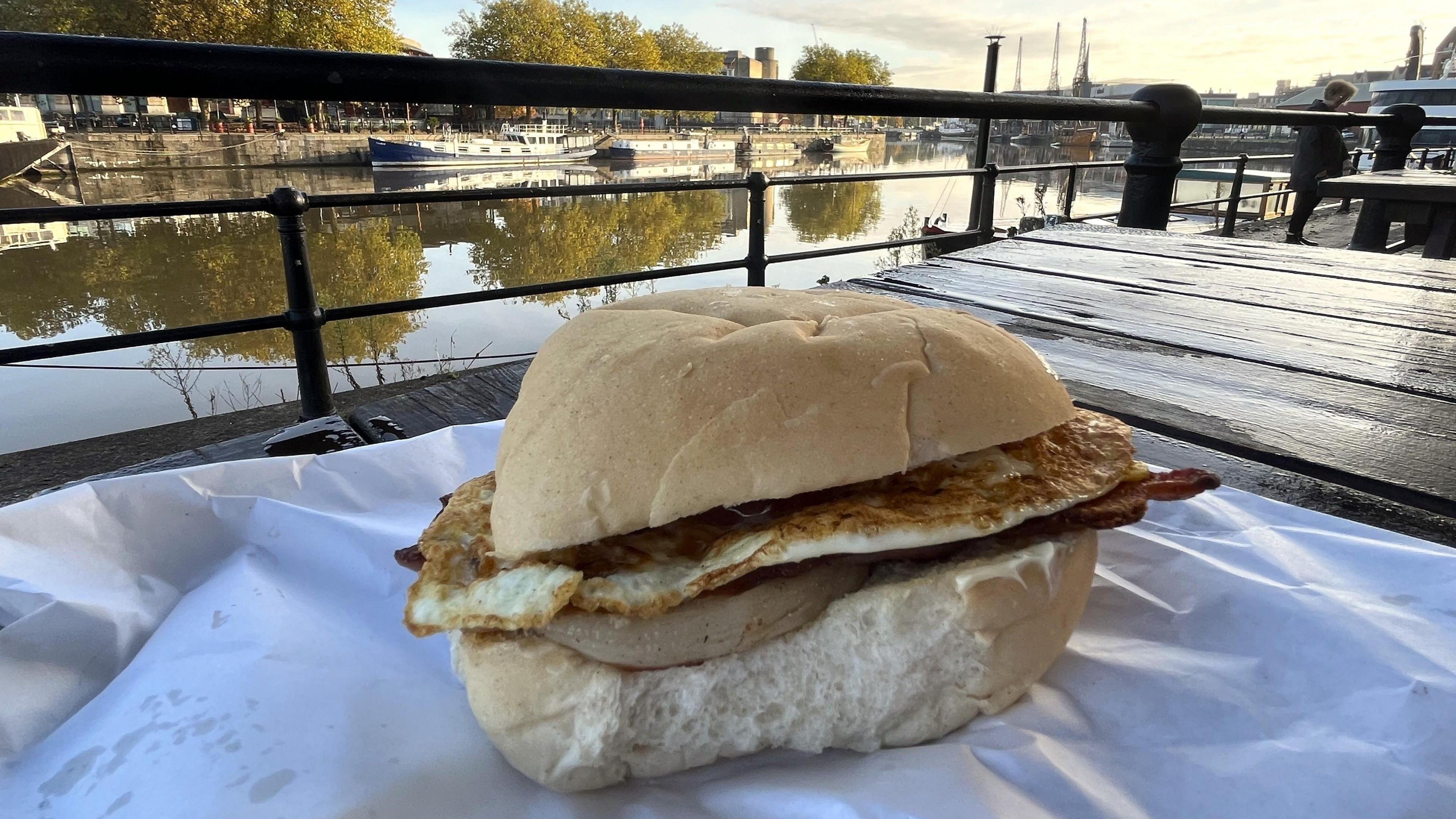 A large egg and bacon bun bought from Bristol's Brunel Buttery is seen sitting on a white paper bag on a table. In the background the city's harbourside can be seen with the MShed cranes visible in the distance