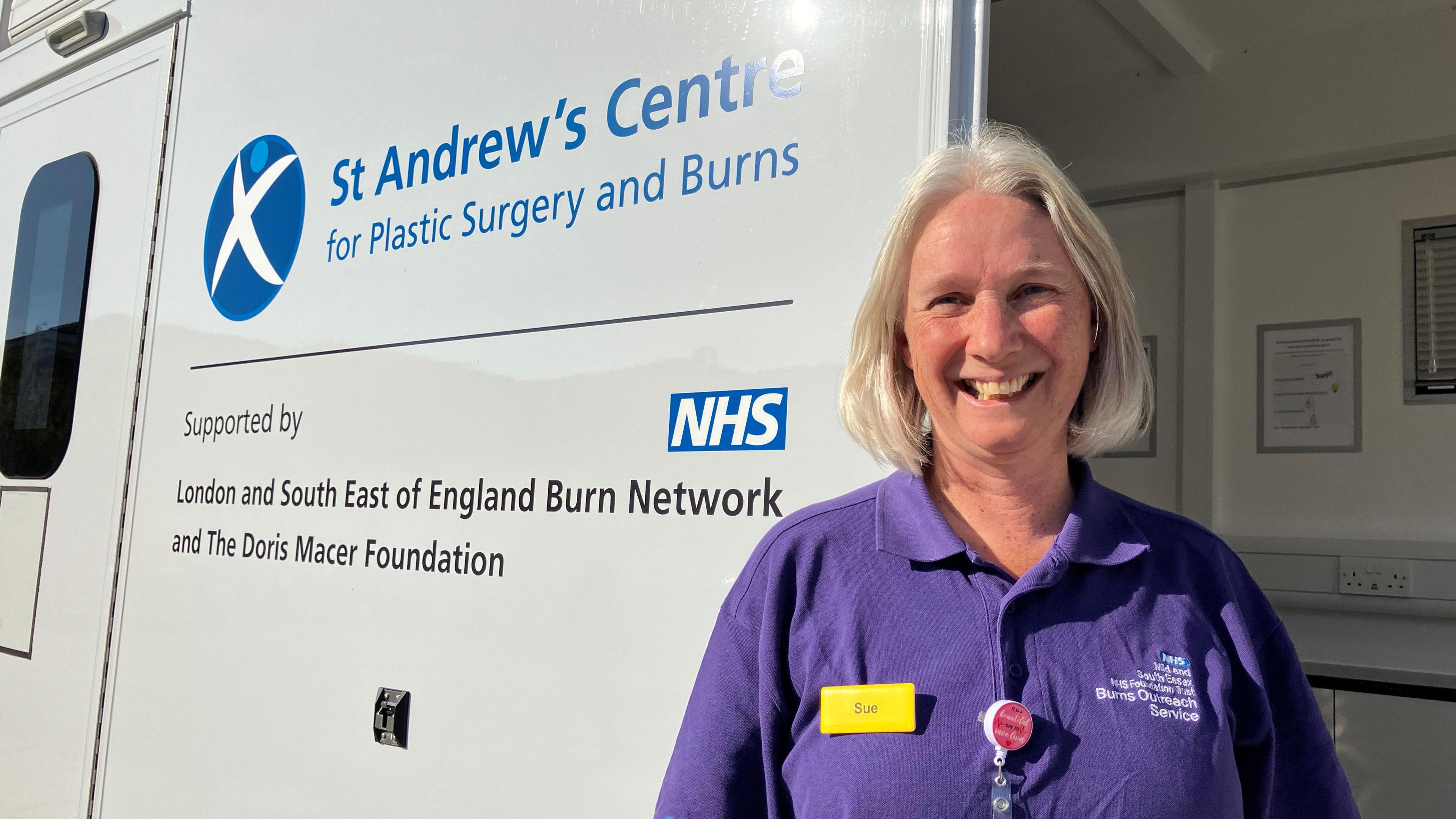 On a sunny day, a woman in a purple top is smiling outside a bus with the words "St Andrew's Centre for plastic surgery and burns" printed on the side of it