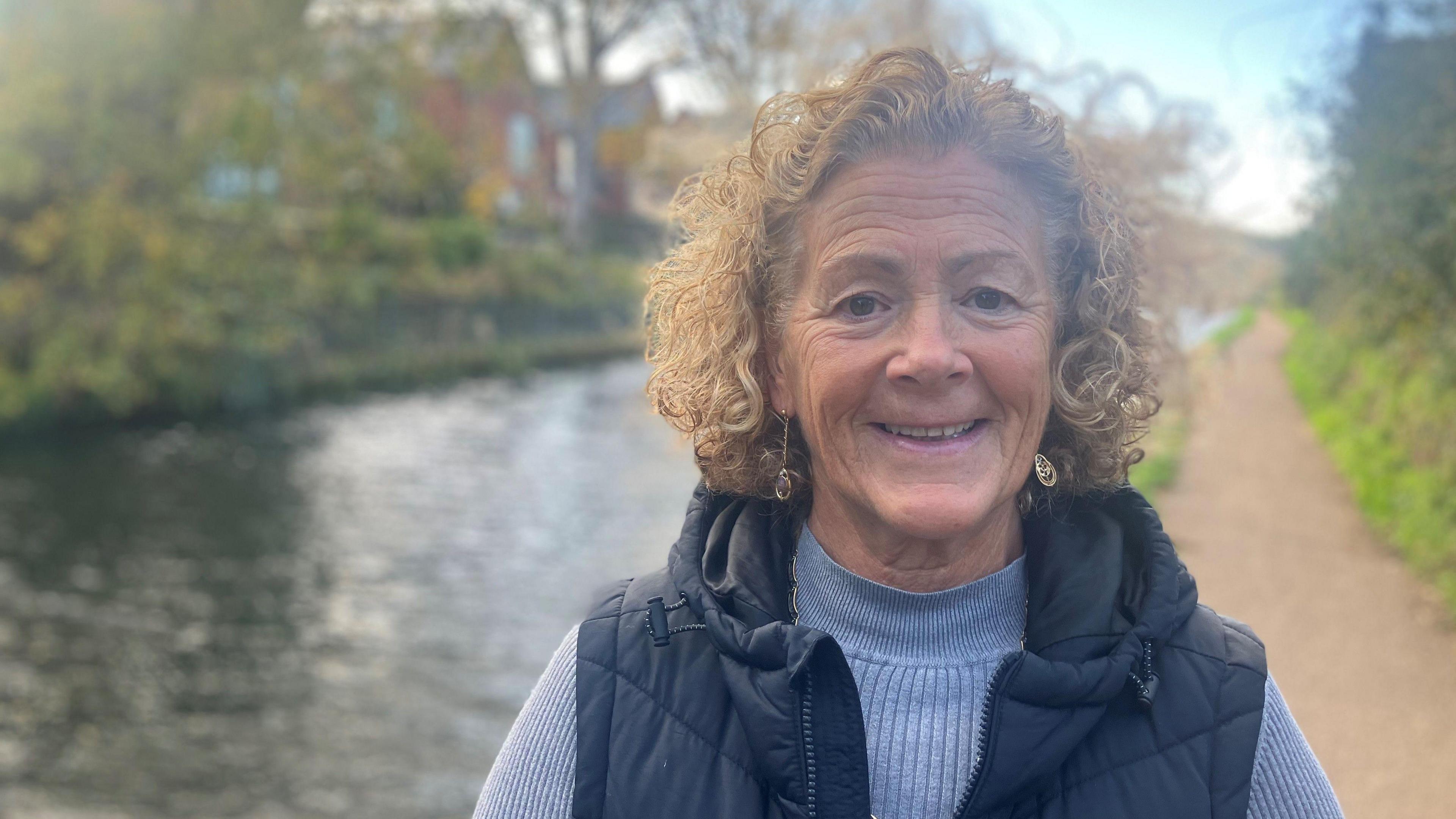 Photograph of Ruth Gallagher, who lives near the proposed Gypsy and traveller site close to Dunham Massey. With curly light brown hair and wearing a navy gilet and light blue round-necked jumper, she is photographed standing on the towpath in front of the Bridgewater Canal.