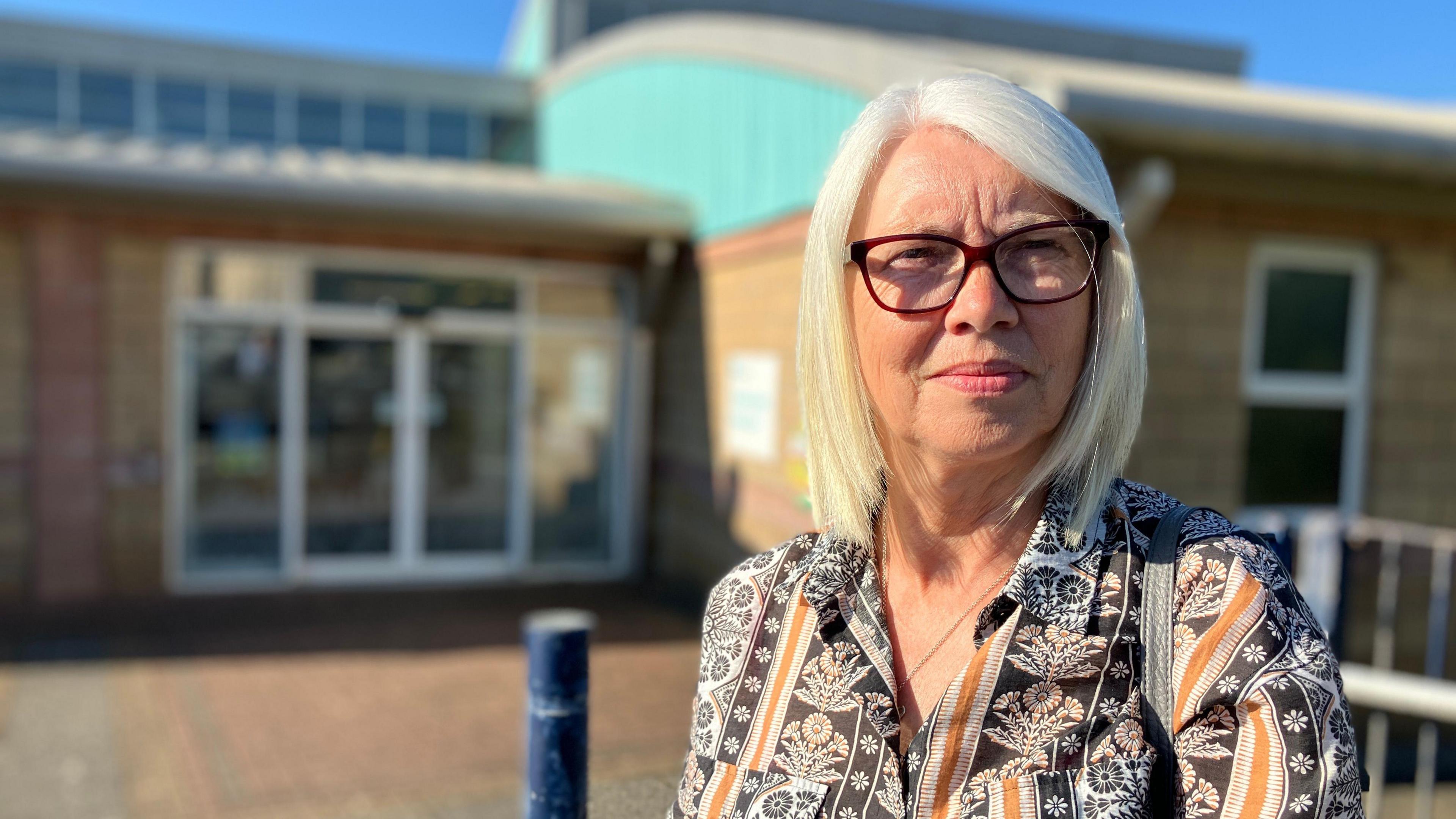 Margaret Heyes, who is blonde and wearing glasses, standing outside the main entrance of the Oval Centre.