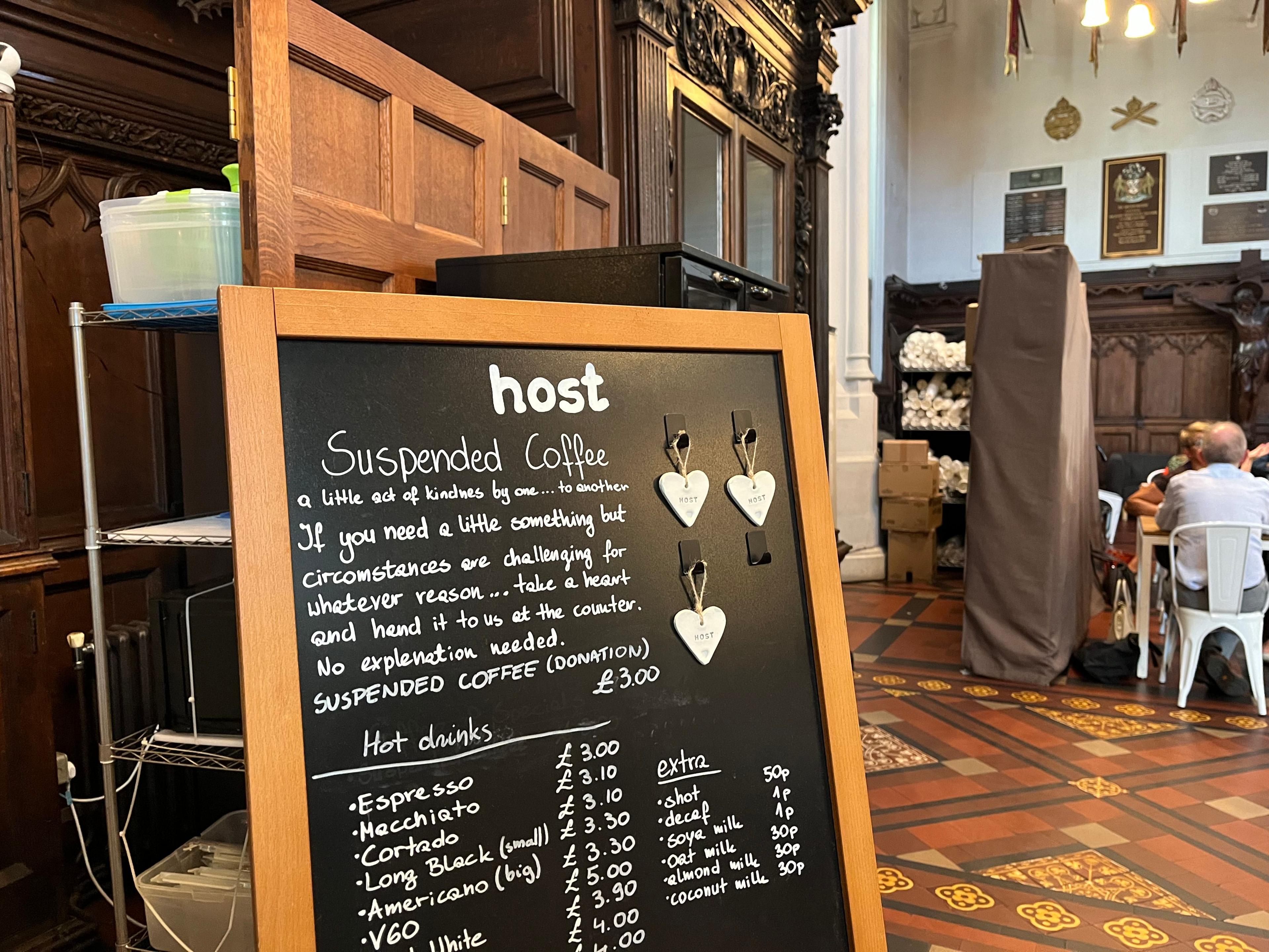 A black chalkboard sign with three hearts hanging off it and a list of drinks and their prices. The top of the sign reads: Suspended Coffee, a little act of kindness by one... to another. 
