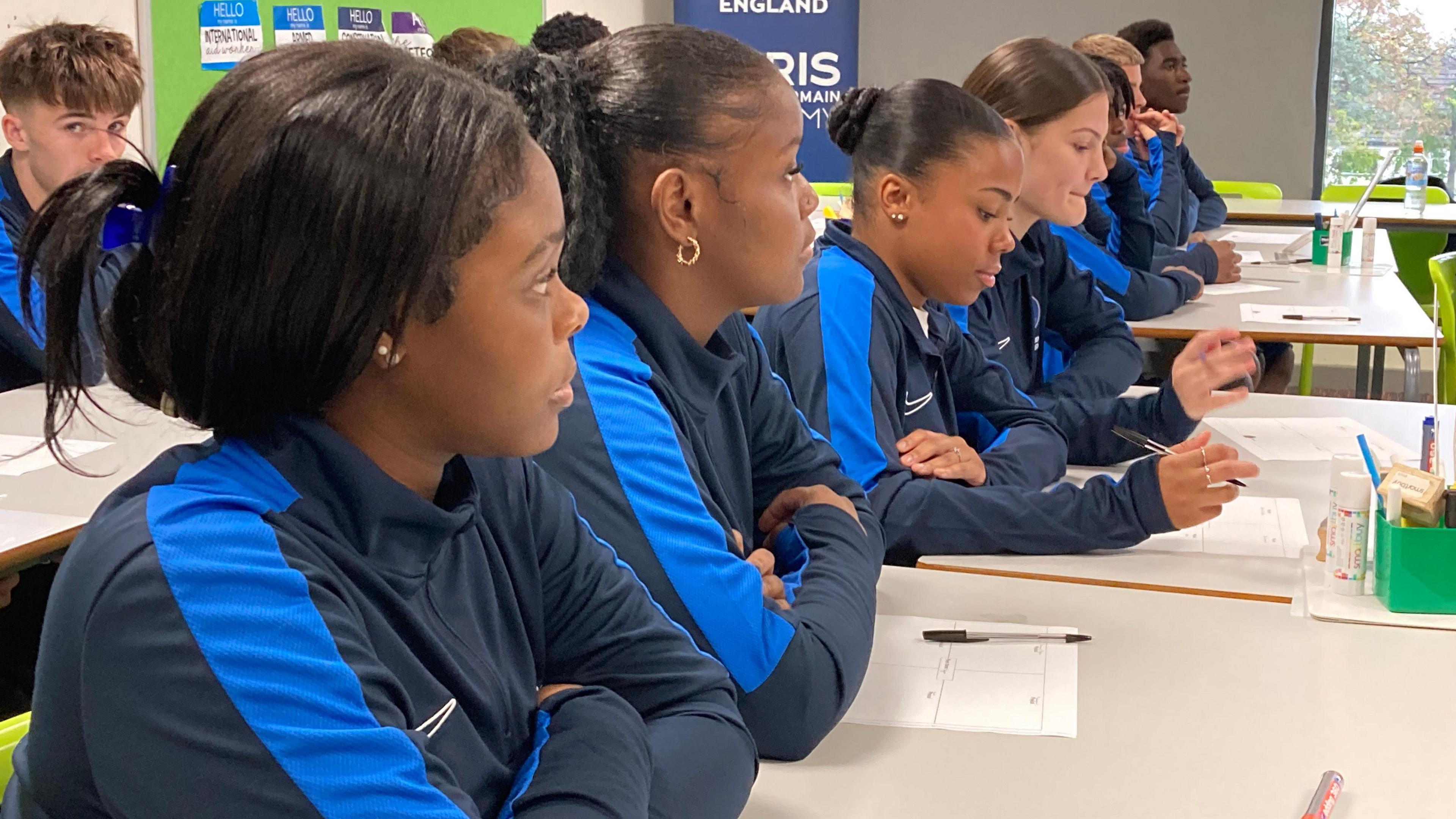 A group of students wearing matching tracksuits sit at desks in a classroom.